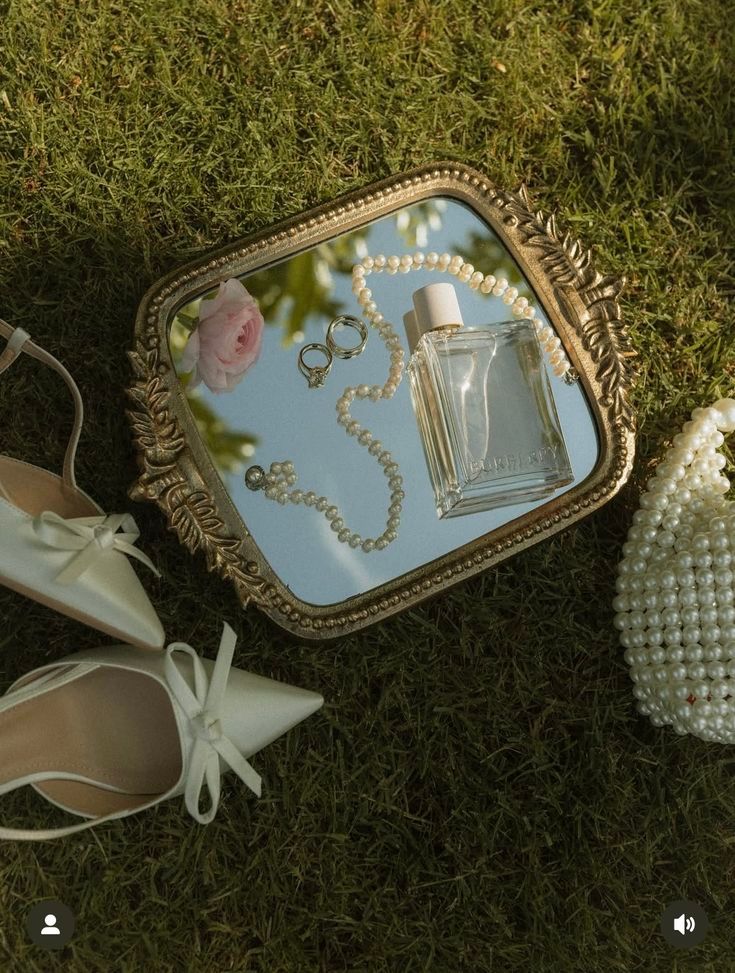 Elegant bridal flat shoes, pearl necklace, perfume bottle, and pearl clutch arranged on a vintage mirror on grass.