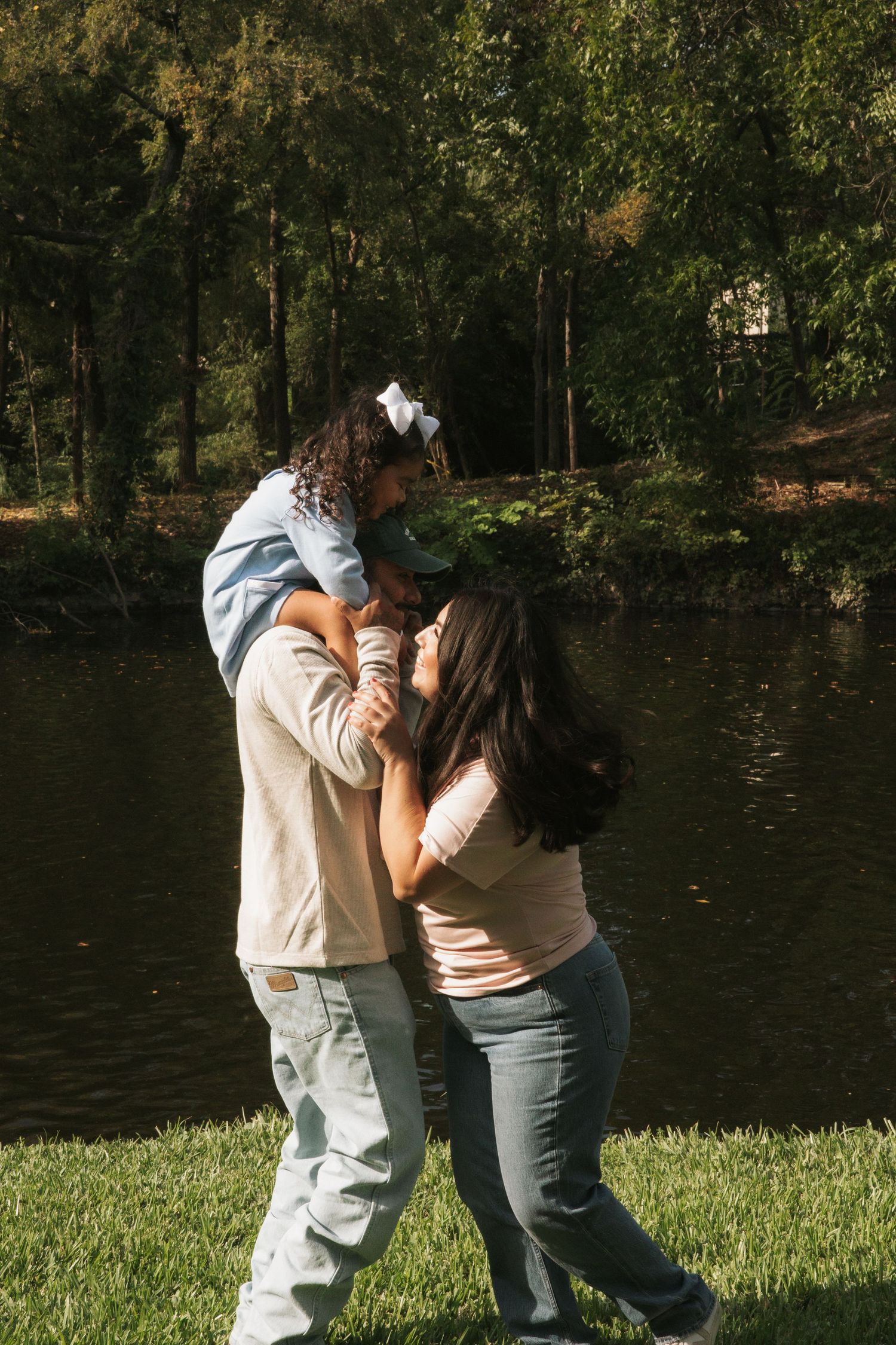 Family sharing a tender moment by a pond, father carrying child on shoulders as mother leans in close.
