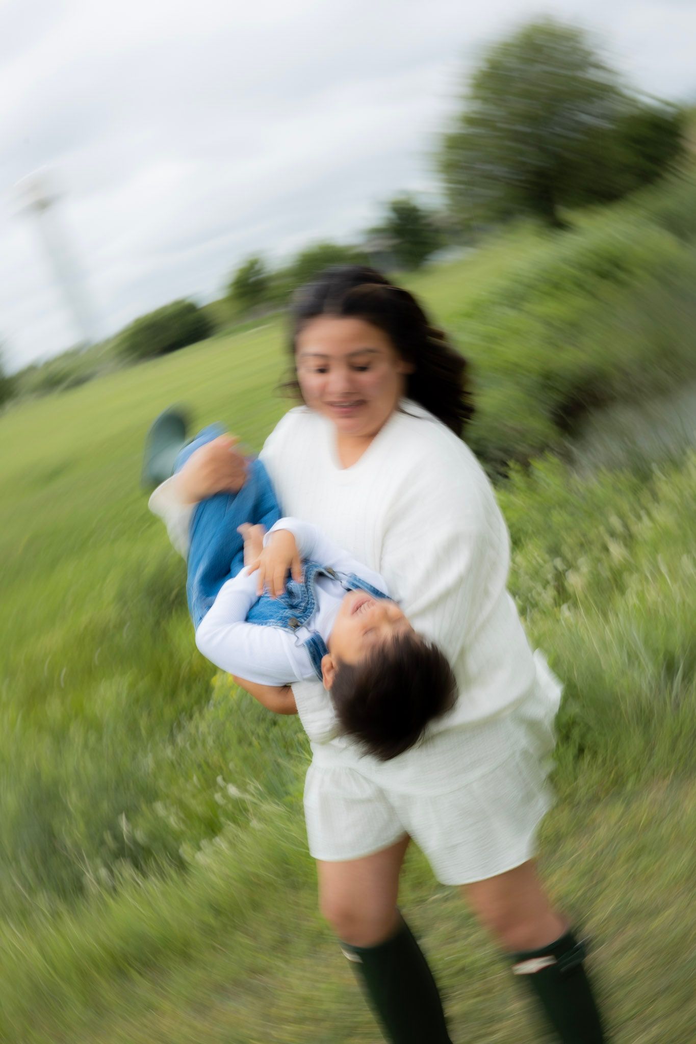 A smiling woman in white spins a laughing child in her arms on a green grassy field outdoors.