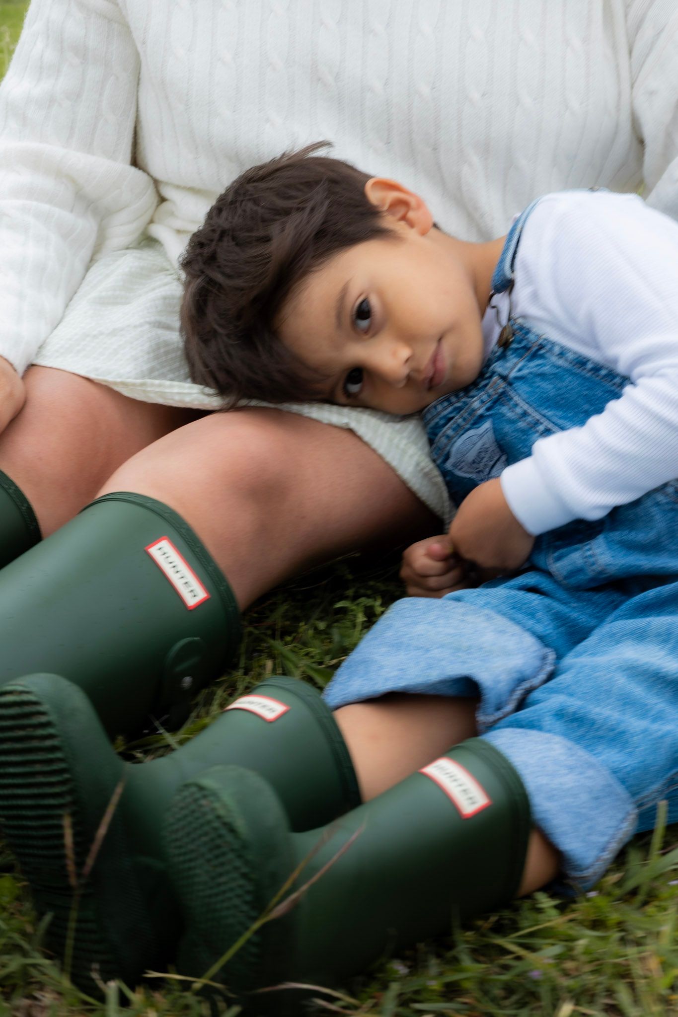 Child in denim overalls rests on adult wearing green Hunter boots on grass.