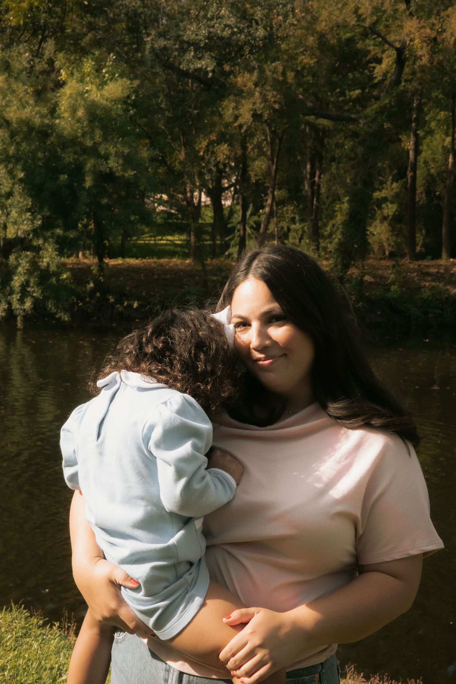 A smiling mother holds her curly-haired child close in a sunny park with a pond and trees in the background.