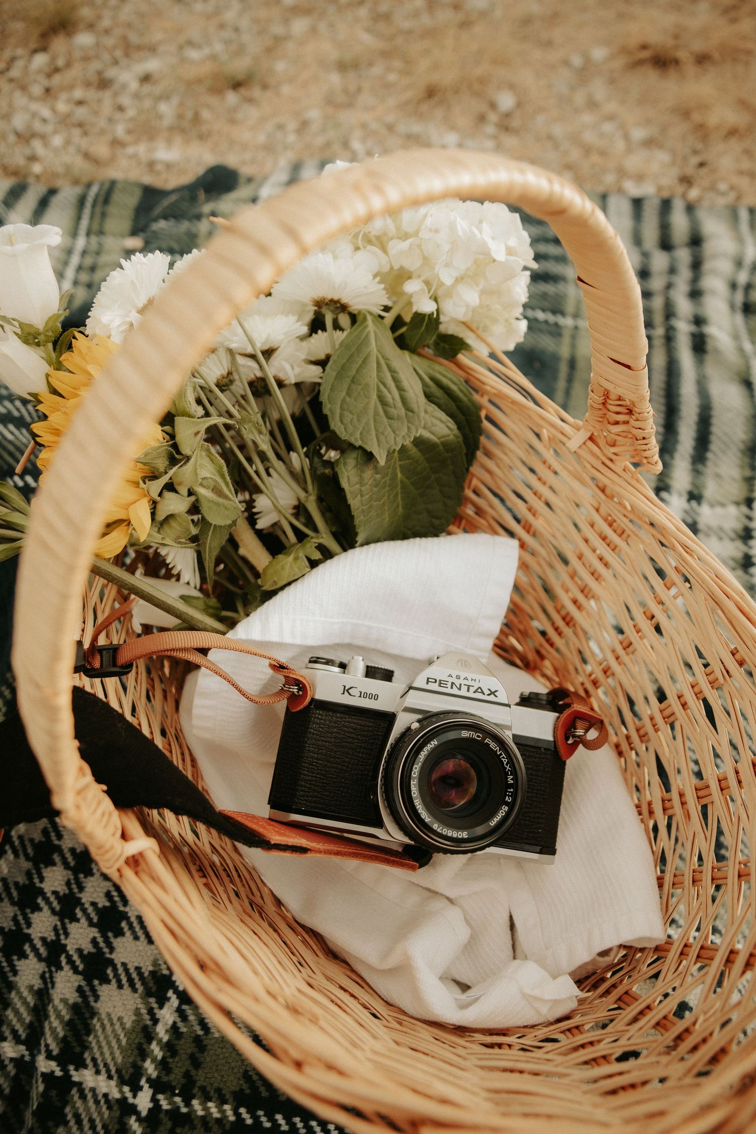 Vintage Pentax camera resting in a wicker picnic basket with white flowers and sunflowers on a plaid blanket.