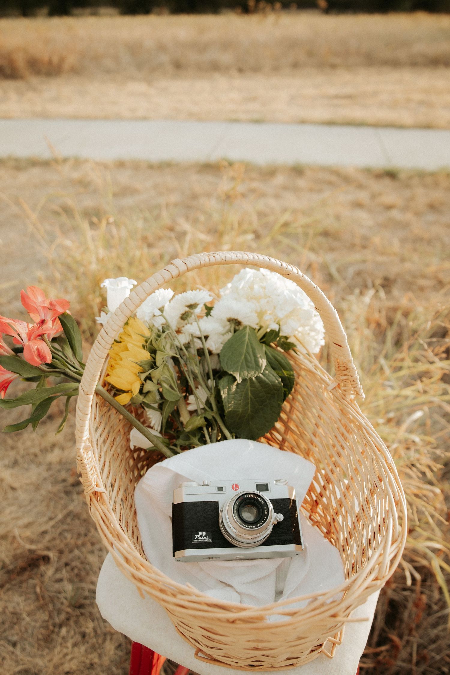 Wicker basket with colorful flowers and vintage film camera resting on white cloth in a sunlit field.