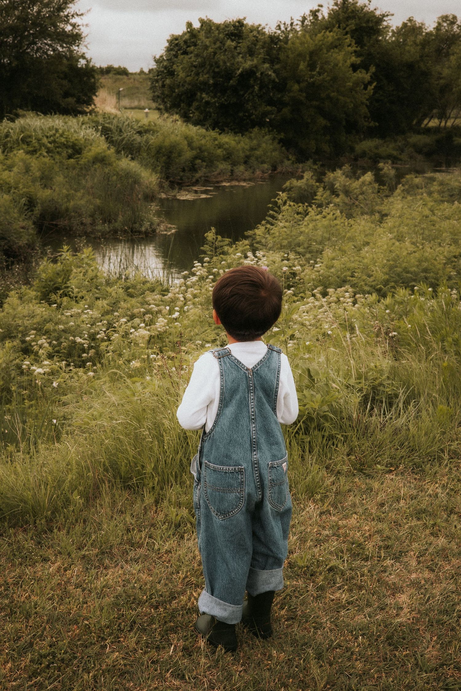 Young child in denim overalls and white shirt stands in lush green field gazing at a serene pond surrounded by wildflowers.