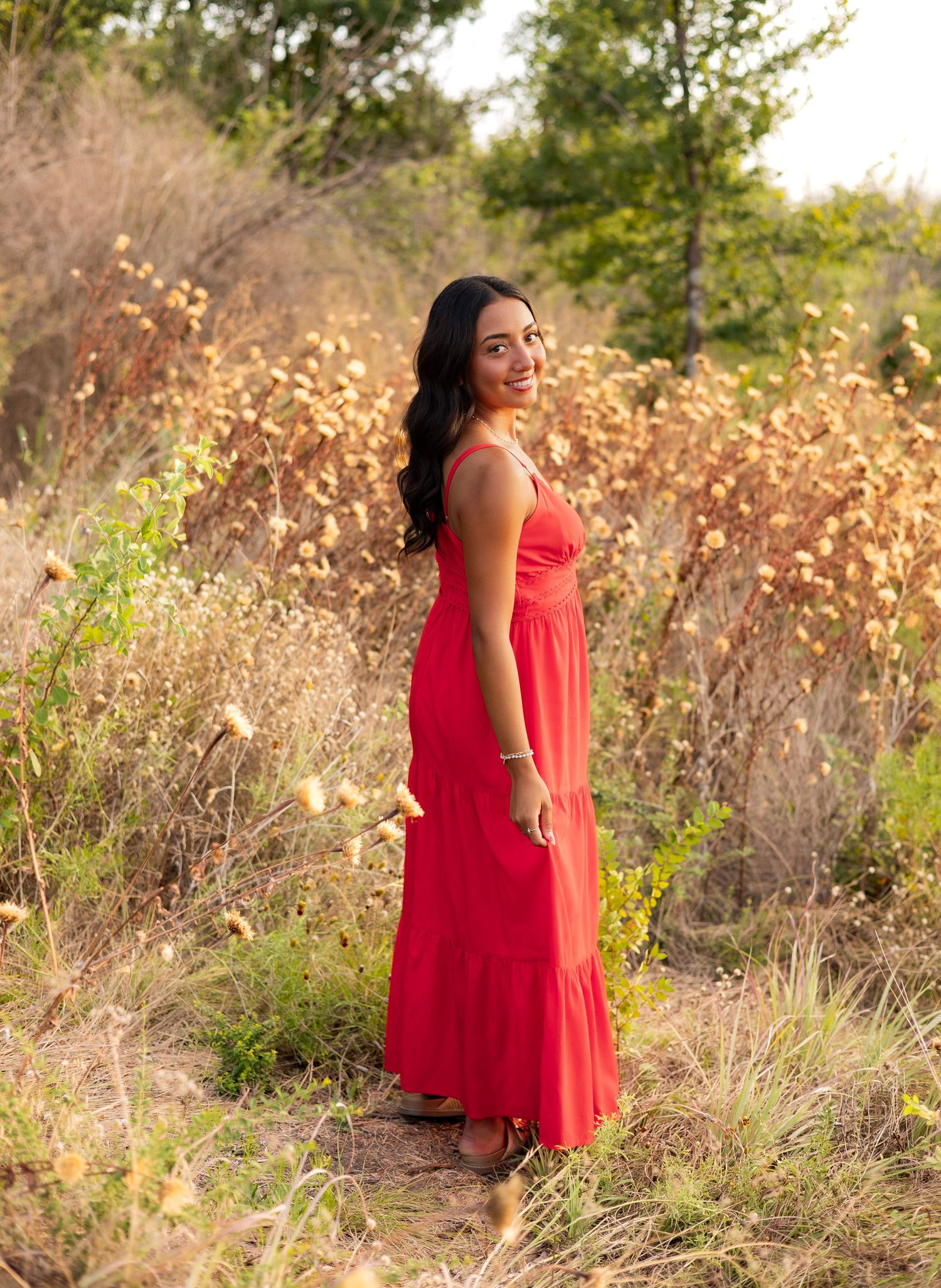 Young woman in red maxi dress standing on a nature trail surrounded by golden wildflowers.