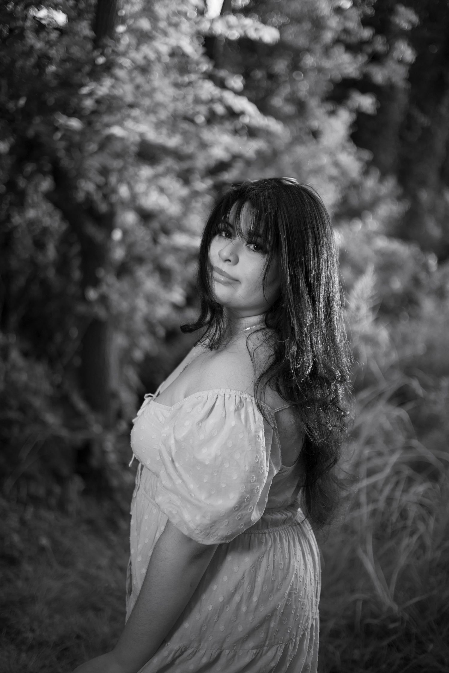 Black and white portrait of young woman in off-shoulder dress standing among trees outdoors.