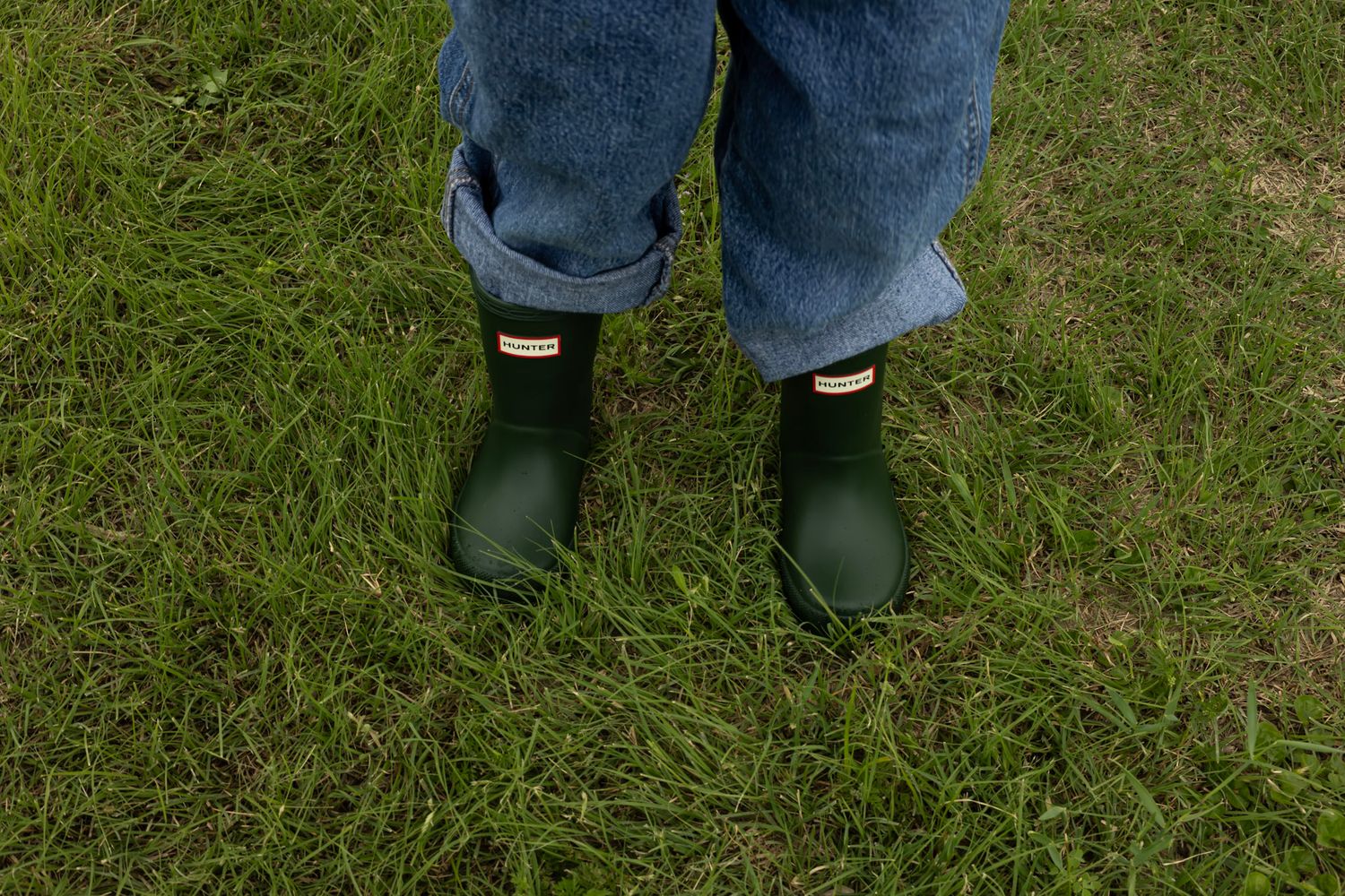 Child wearing green Hunter rain boots and rolled-up jeans standing on lush green grass.