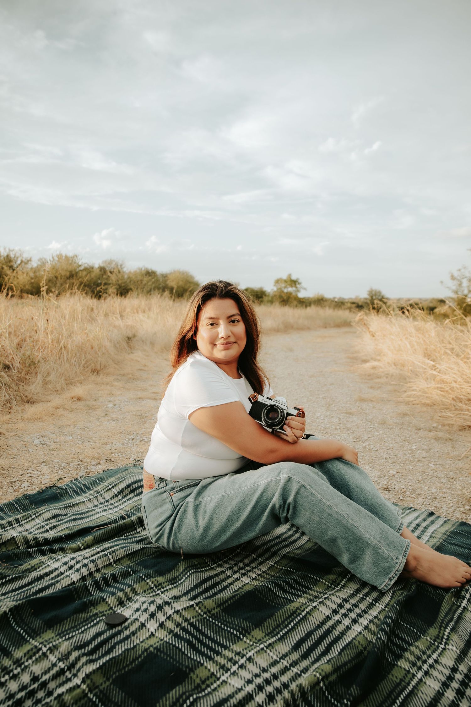 Young woman sitting on plaid blanket outdoors holding camera, smiling in natural field setting.