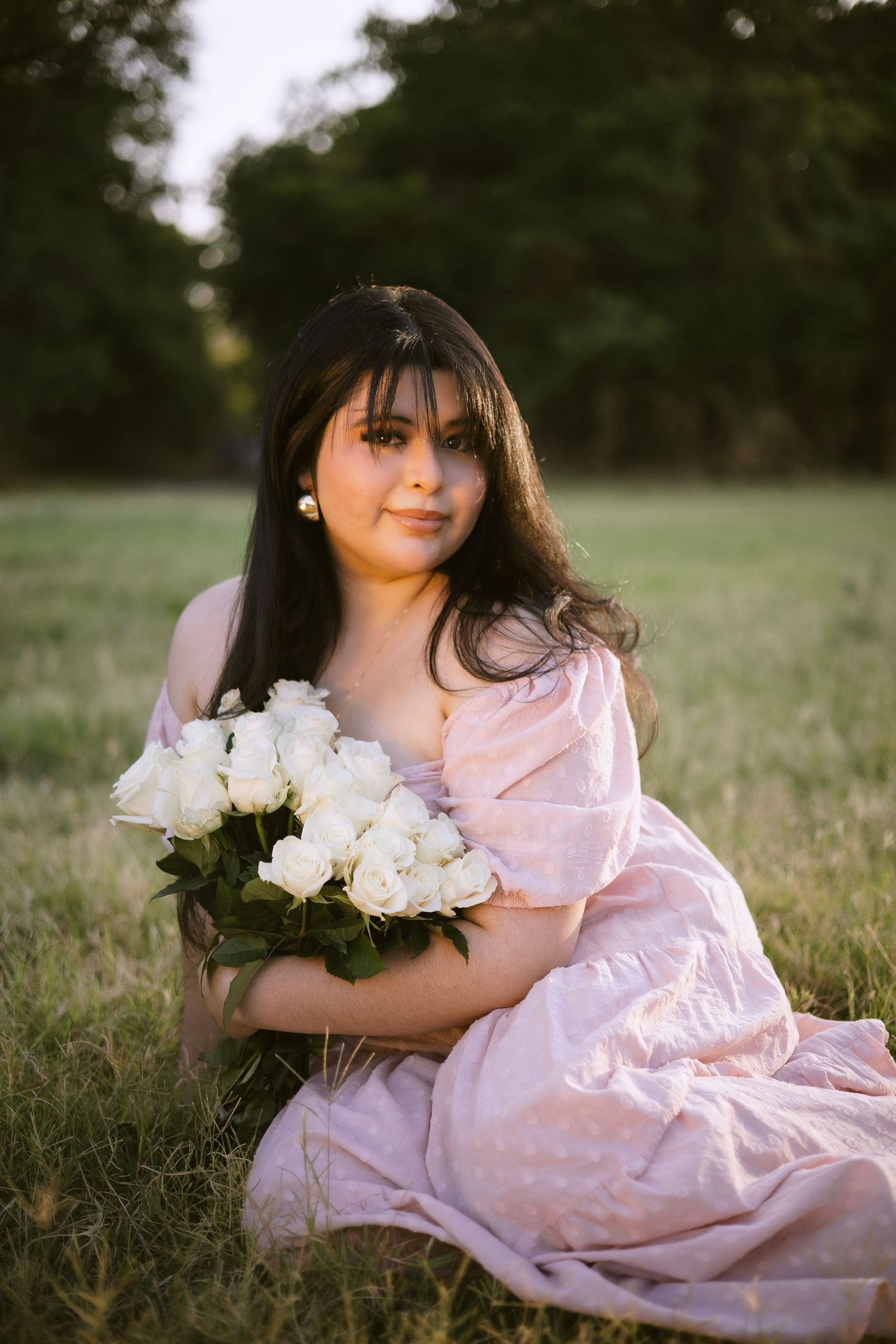 Young woman in pink puff-sleeve dress hugging white roses bouquet while sitting in golden grass.