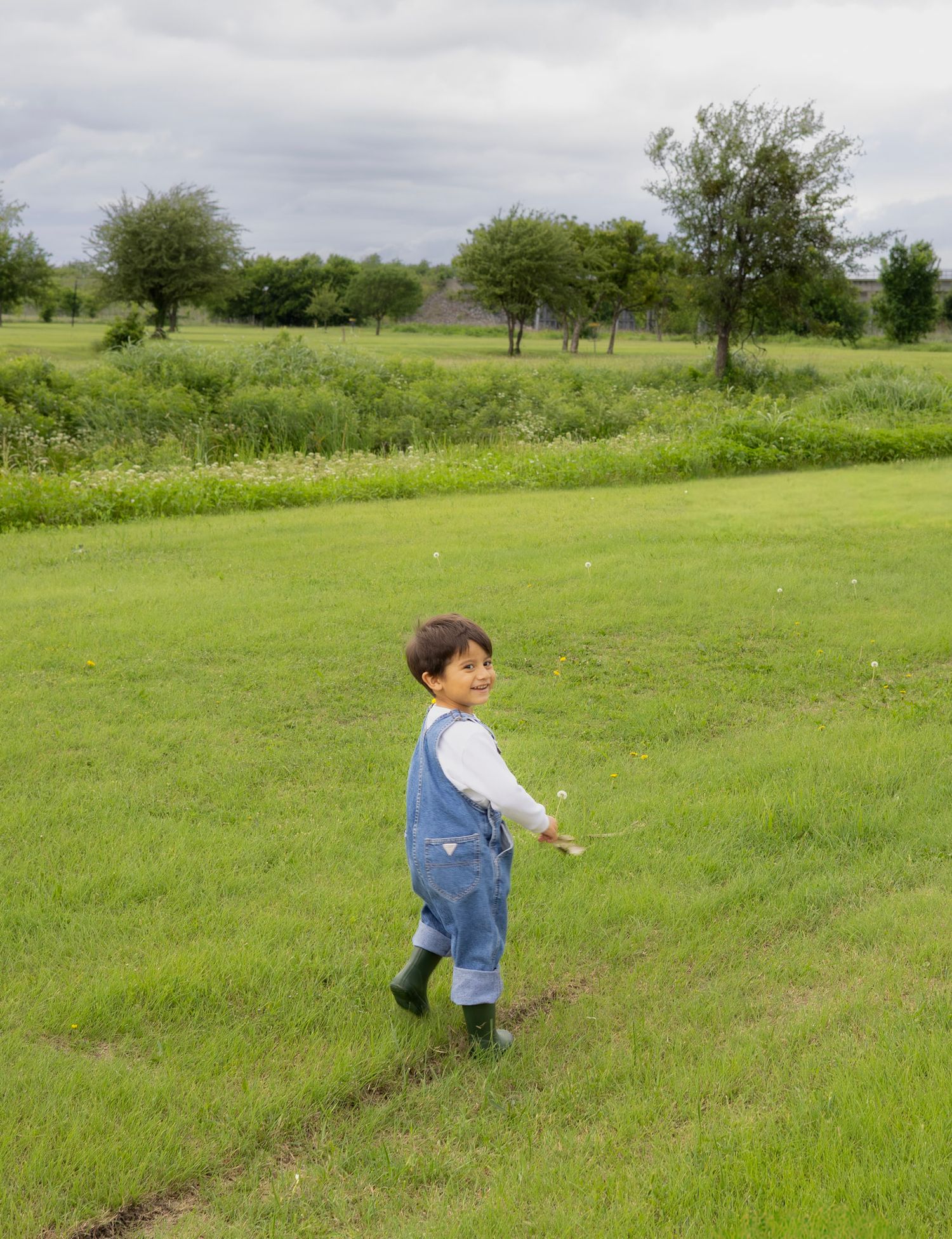 A happy toddler in denim overalls and green Hunter boots walks across a lush green grassy field.