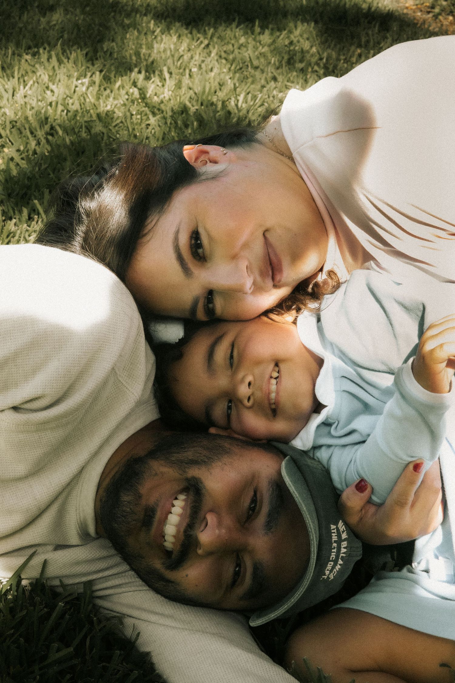 Smiling family of three lying on grass together looking up at camera in a sunlit park.