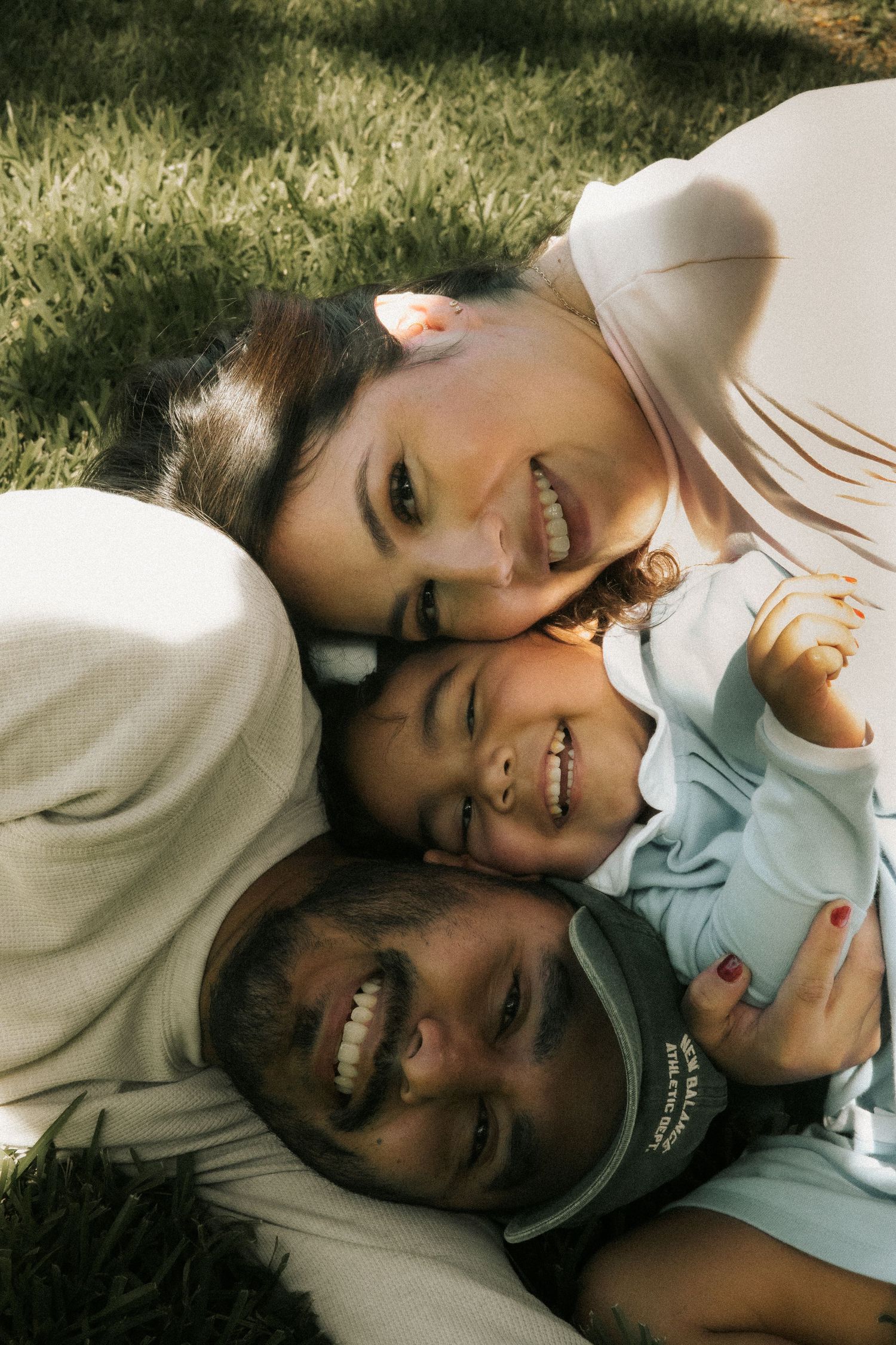Happy family of three lying on grass, smiling together in a warm outdoor portrait.