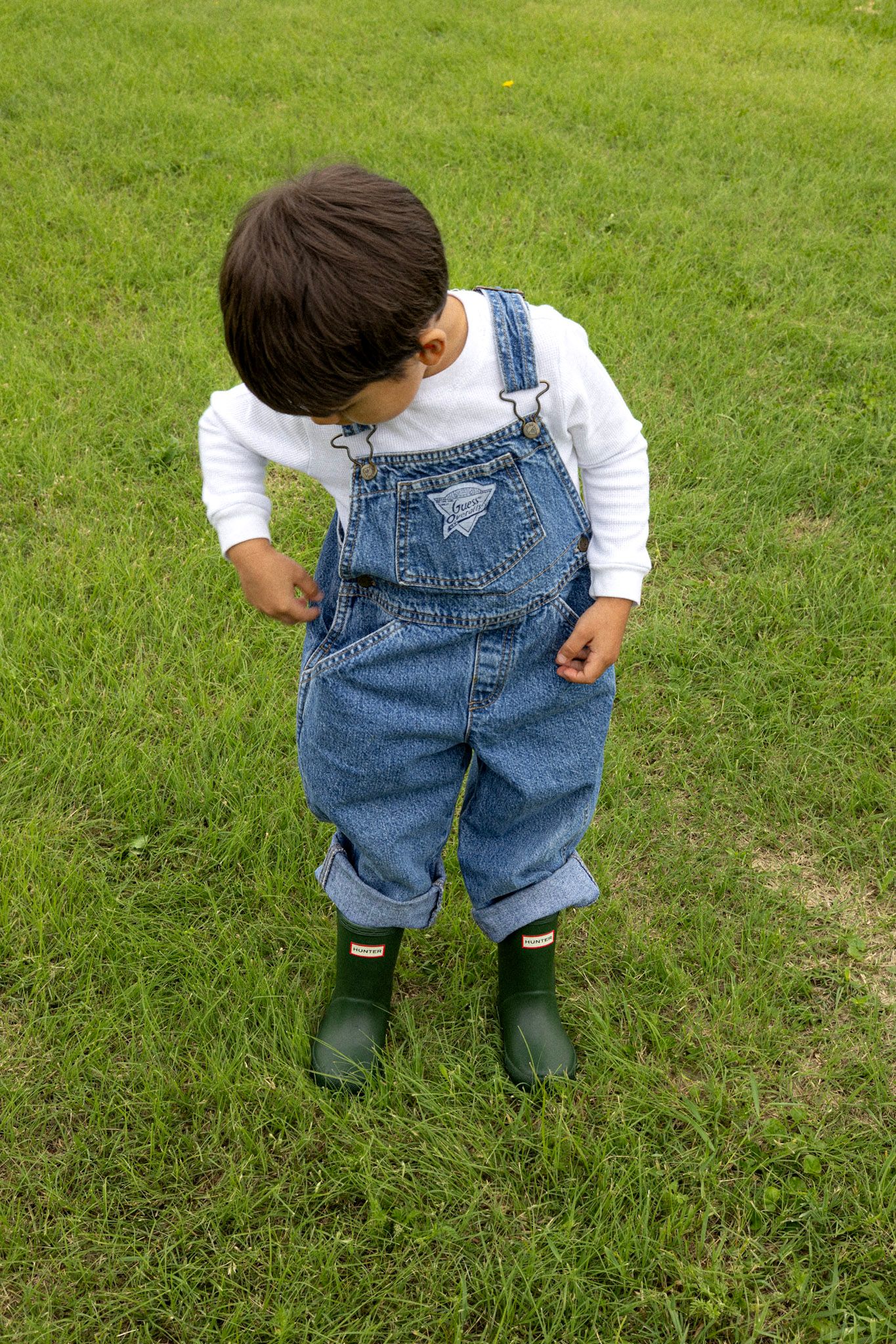 Toddler in denim overalls and green rain boots standing on green grass looking down.