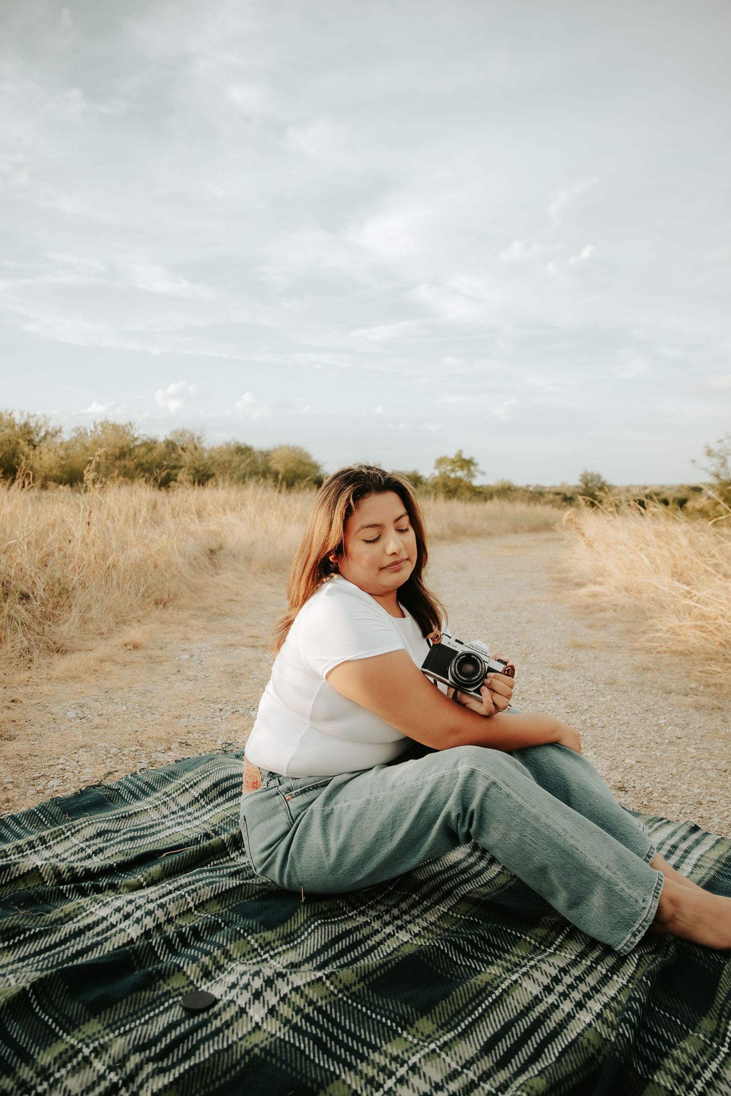 Woman sitting on plaid blanket outdoors holding vintage camera, wearing white tee and jeans, surrounded by dry grass fields.
