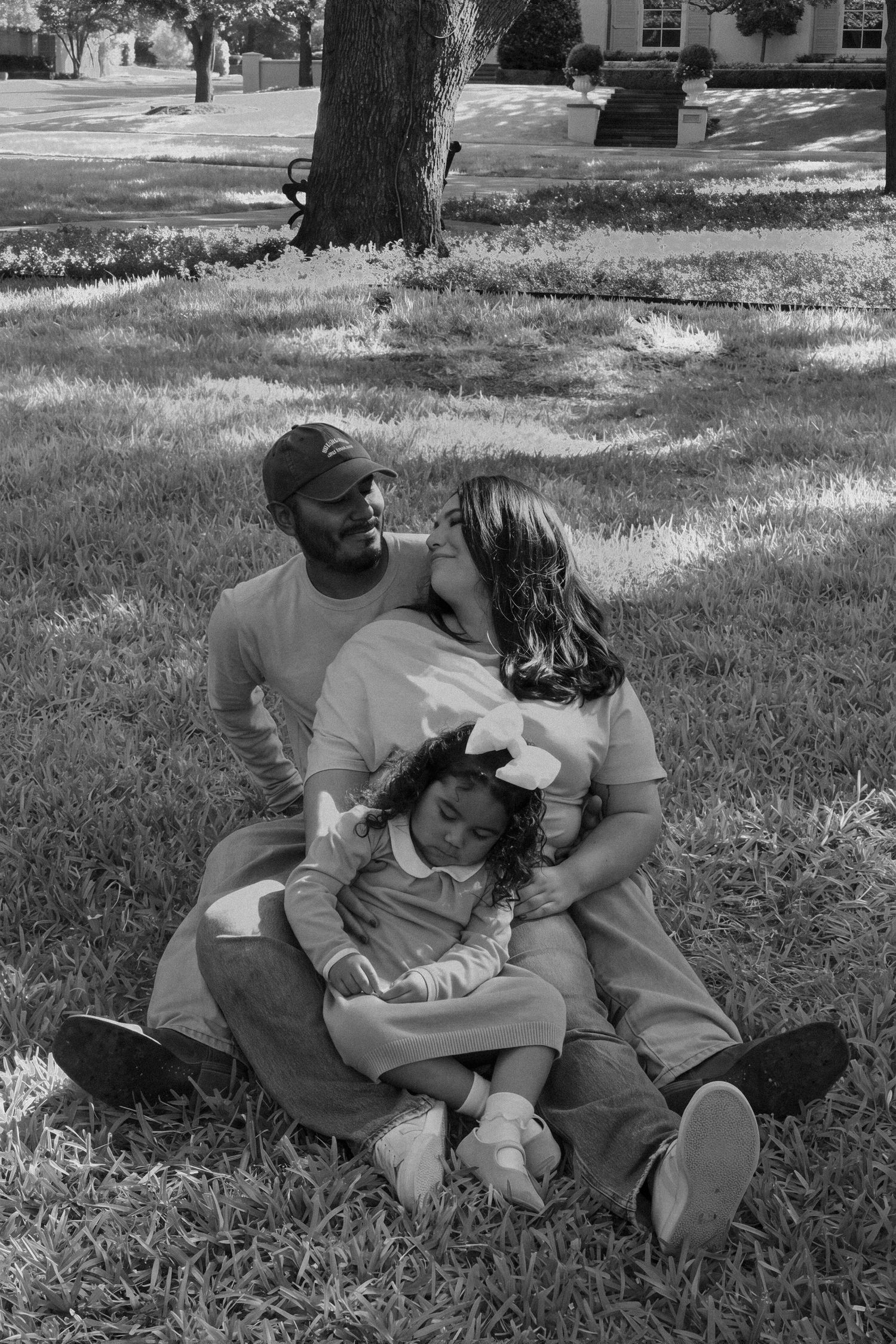 Black and white photo of loving family of three sitting together on grass in a sunlit park.
