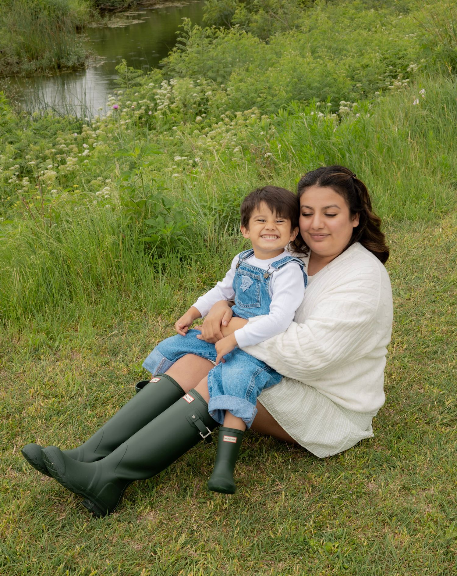A mother and toddler sit together on green grass, both smiling, wearing denim outfits and green rain boots near a pond.