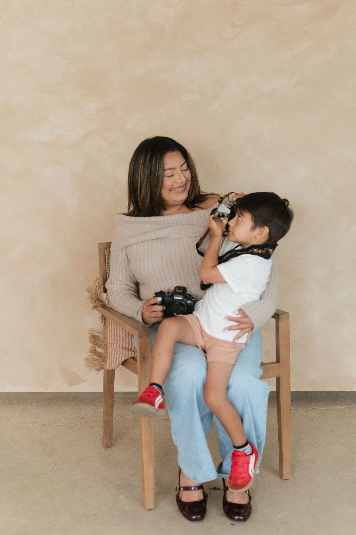 A woman sits on a wooden chair holding a camera while a young child stands on her lap looking through a camera lens.