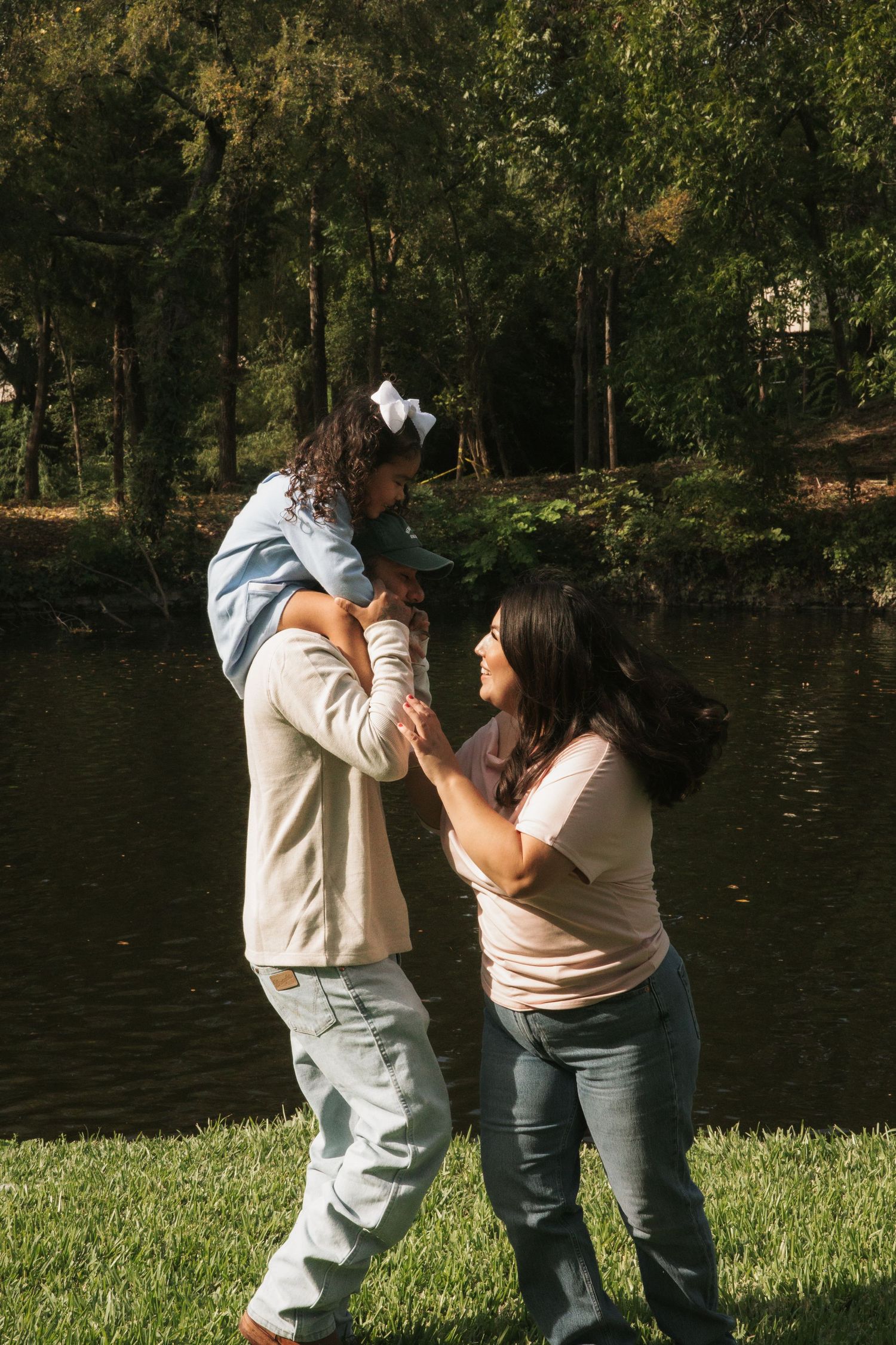 Parents playfully lifting their young daughter by a peaceful lakeside in a sunny park.