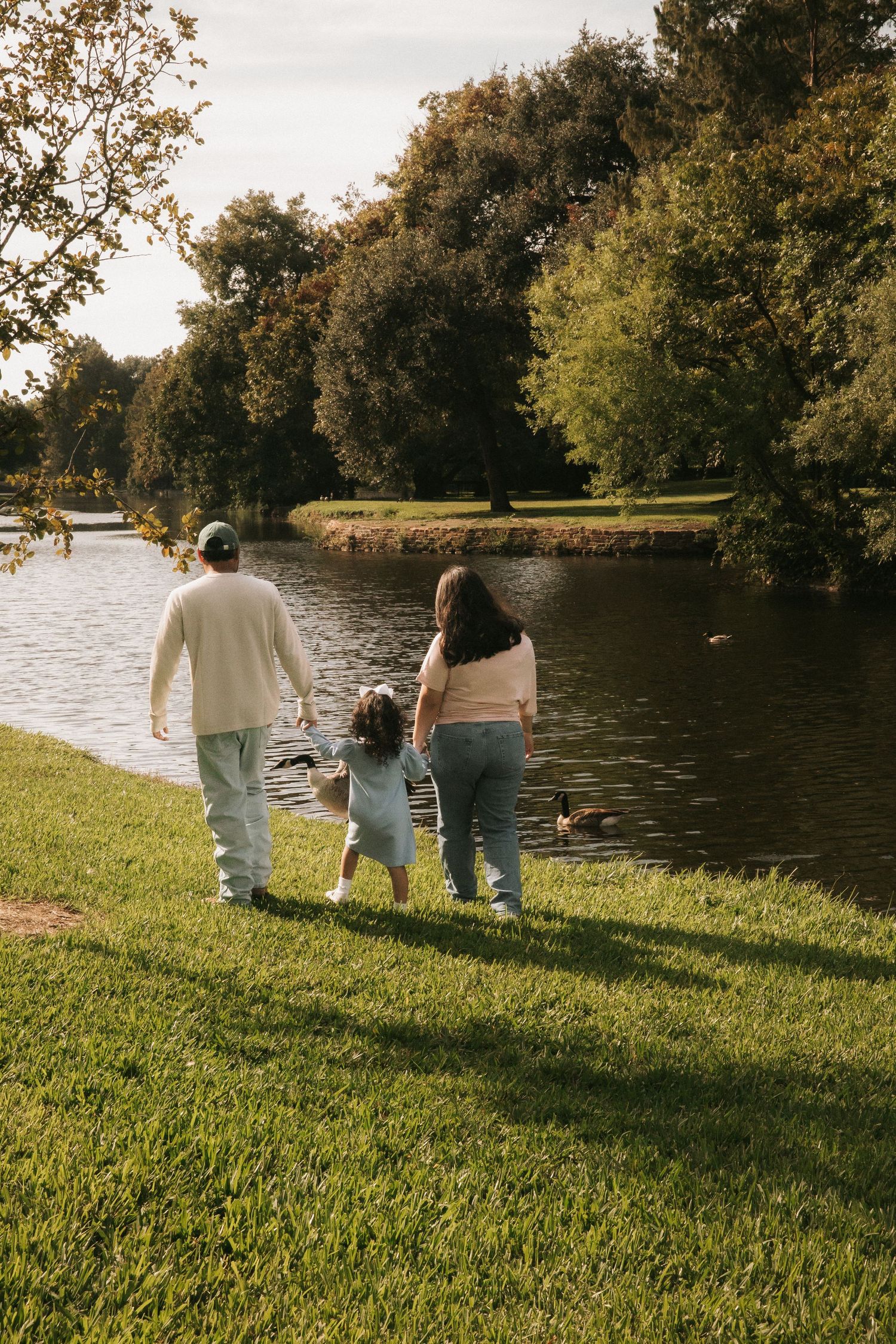 Family of three walking hand-in-hand toward a serene pond in a lush green park on a sunny day.