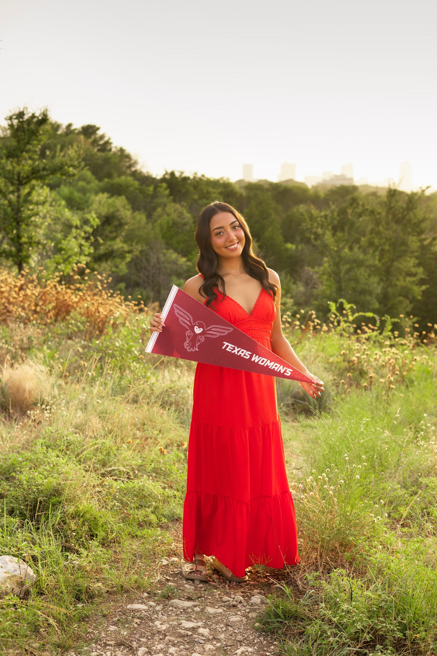Young woman in red dress holding Texas Womans University pennant in a sunlit outdoor field.