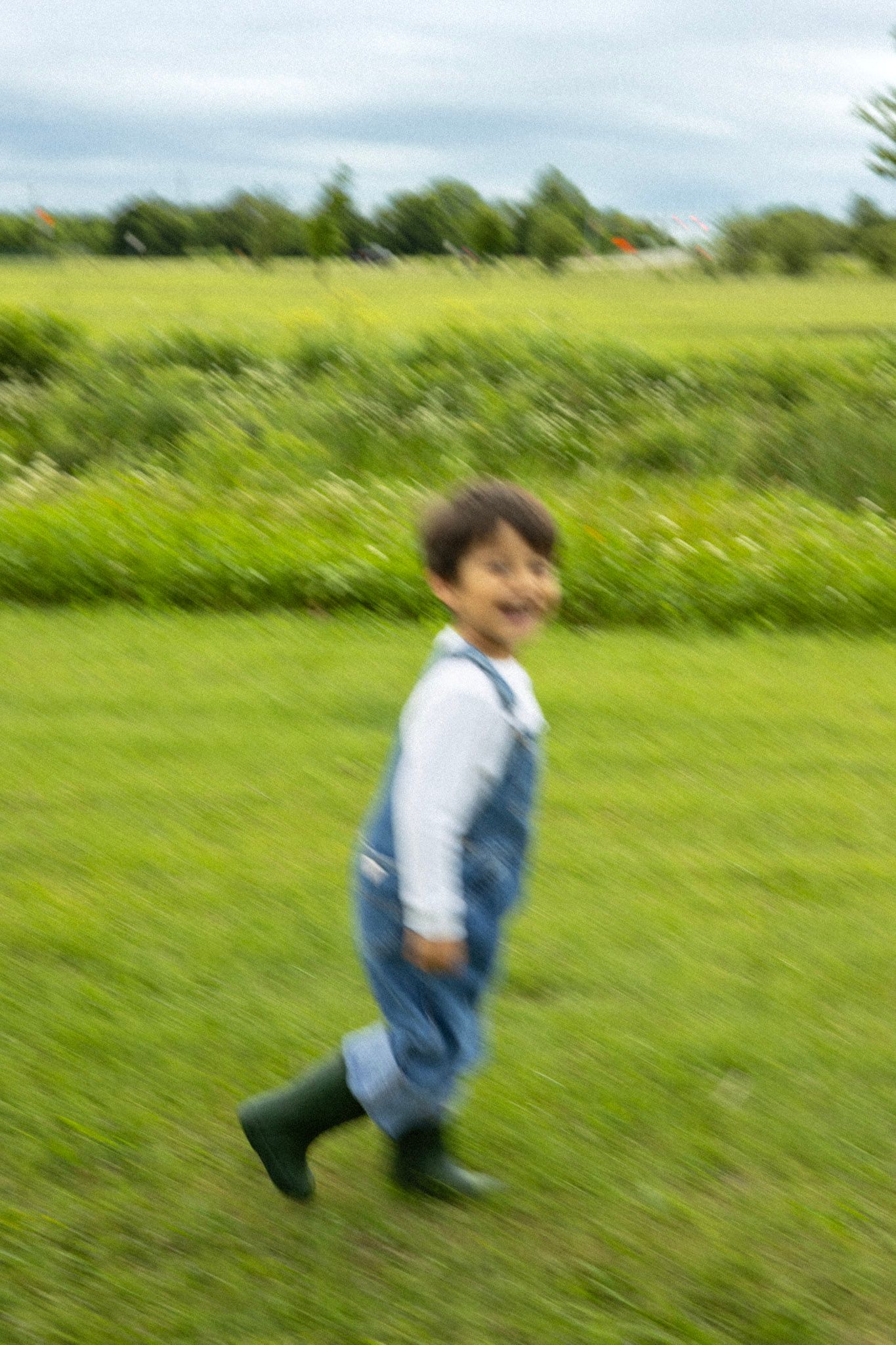 Happy toddler running in denim overalls and green boots across a green field, motion blur.