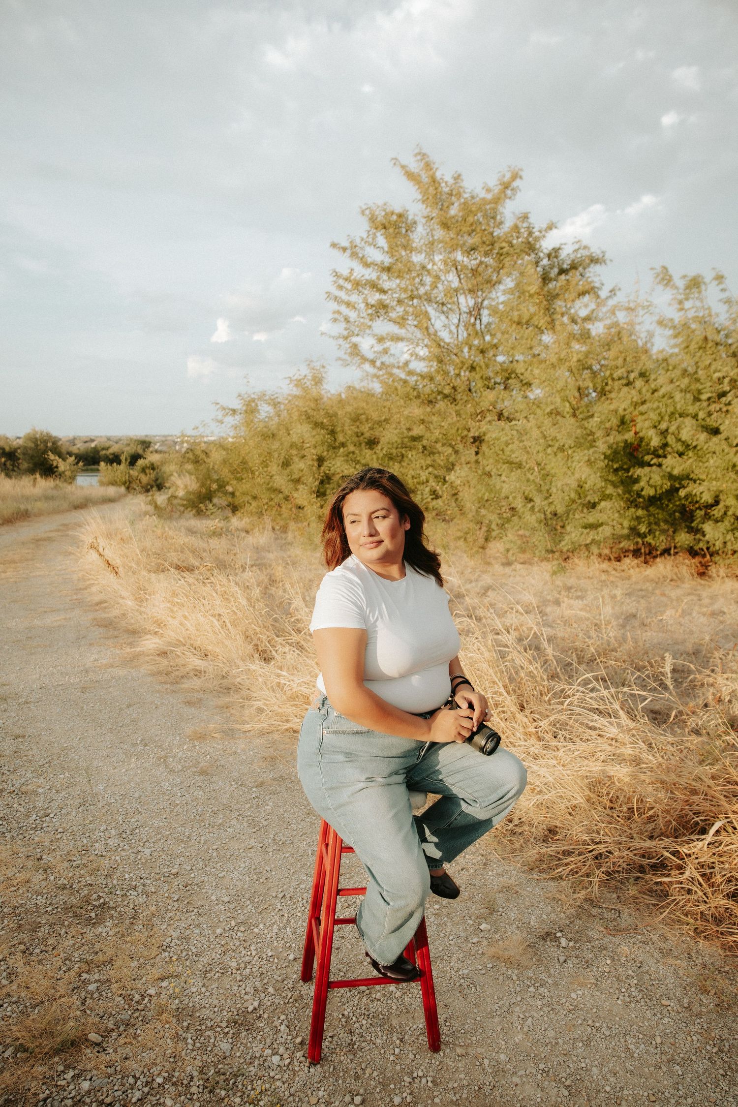 Young woman in white tee and jeans sits on red stool on rural dirt path surrounded by dry grass and trees.