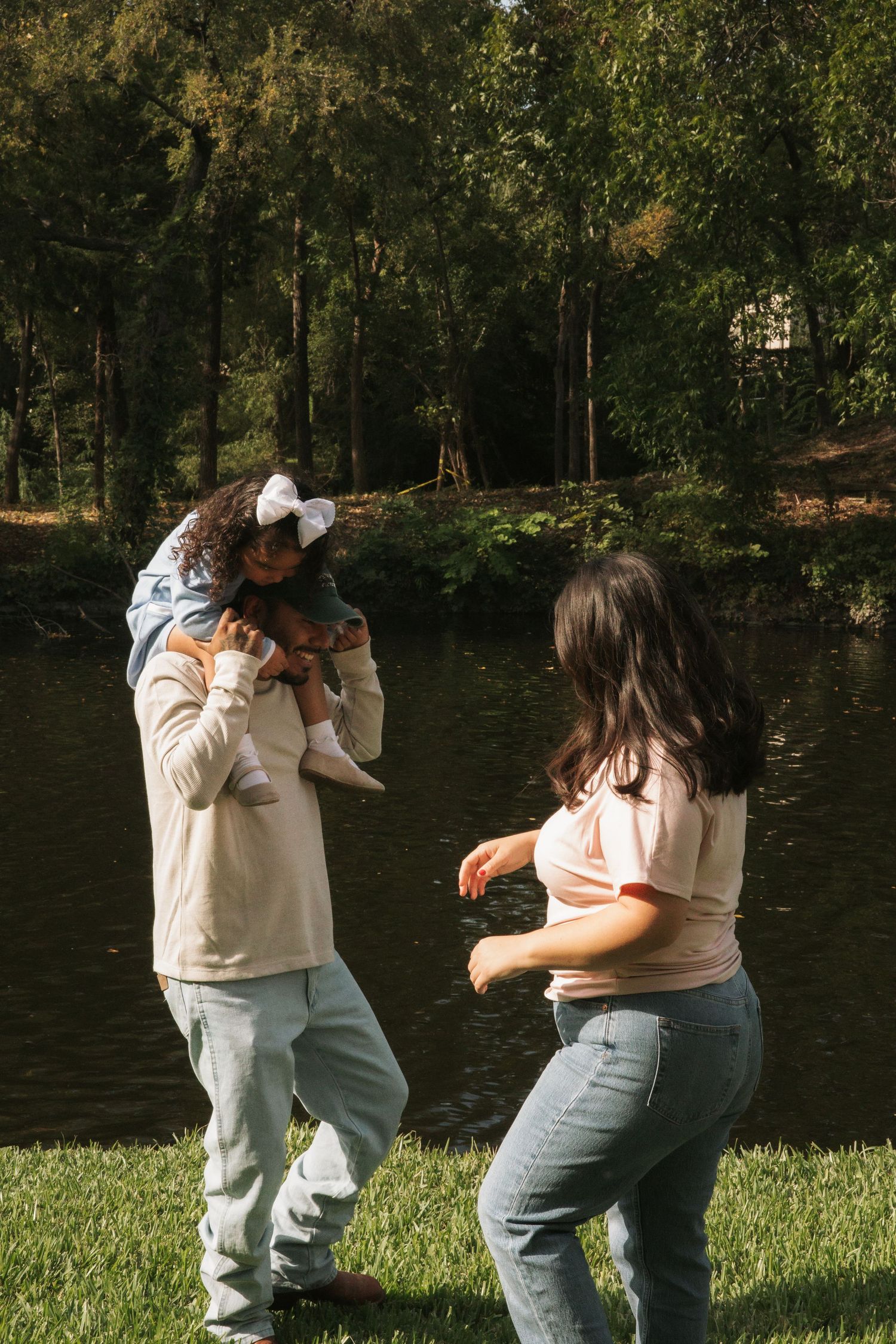 Family playing outdoors near a pond, with a child on shoulders, surrounded by lush green trees on a sunny day.