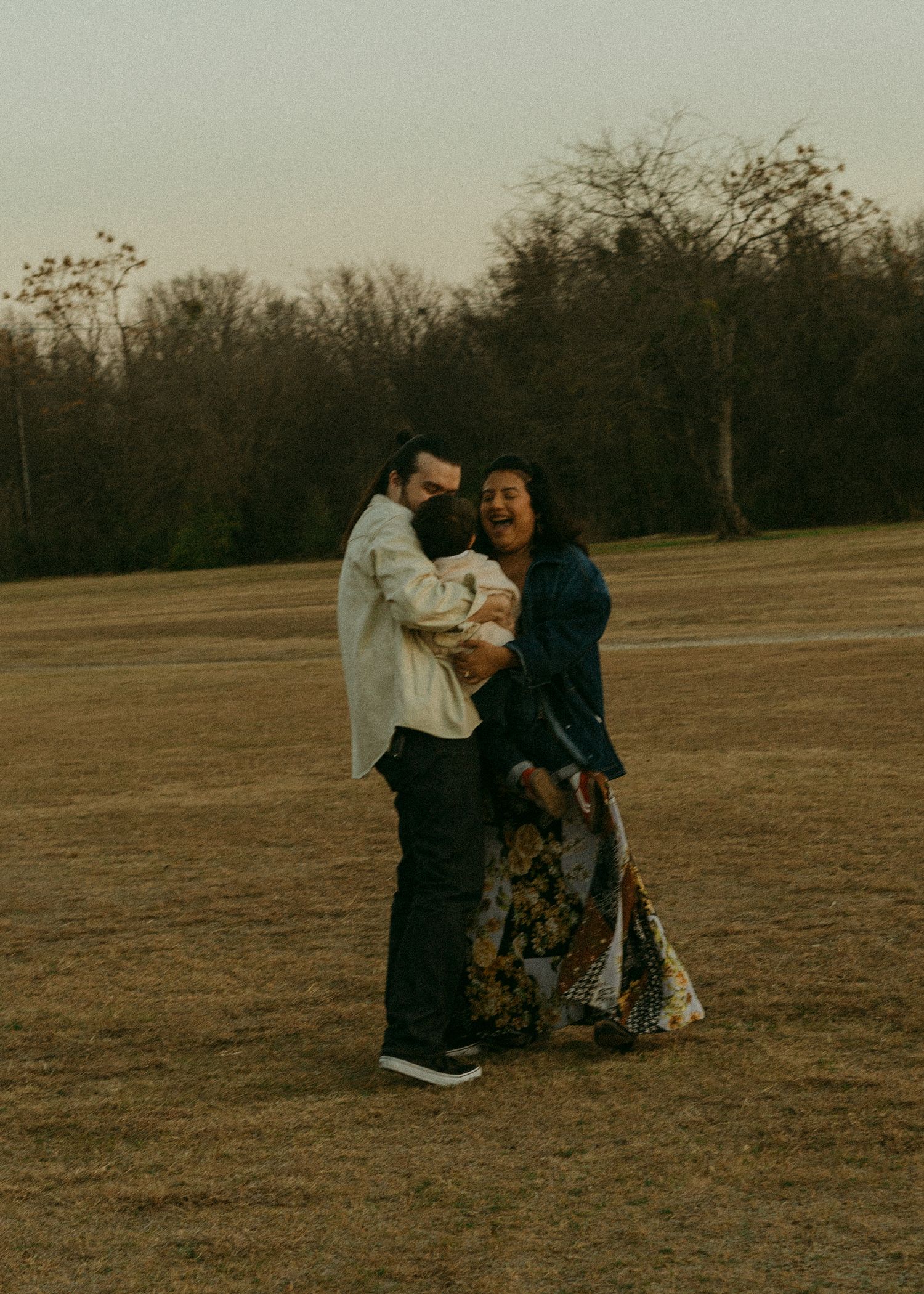 A joyful family of three embraces and laughs together in an open field at dusk, surrounded by bare trees.