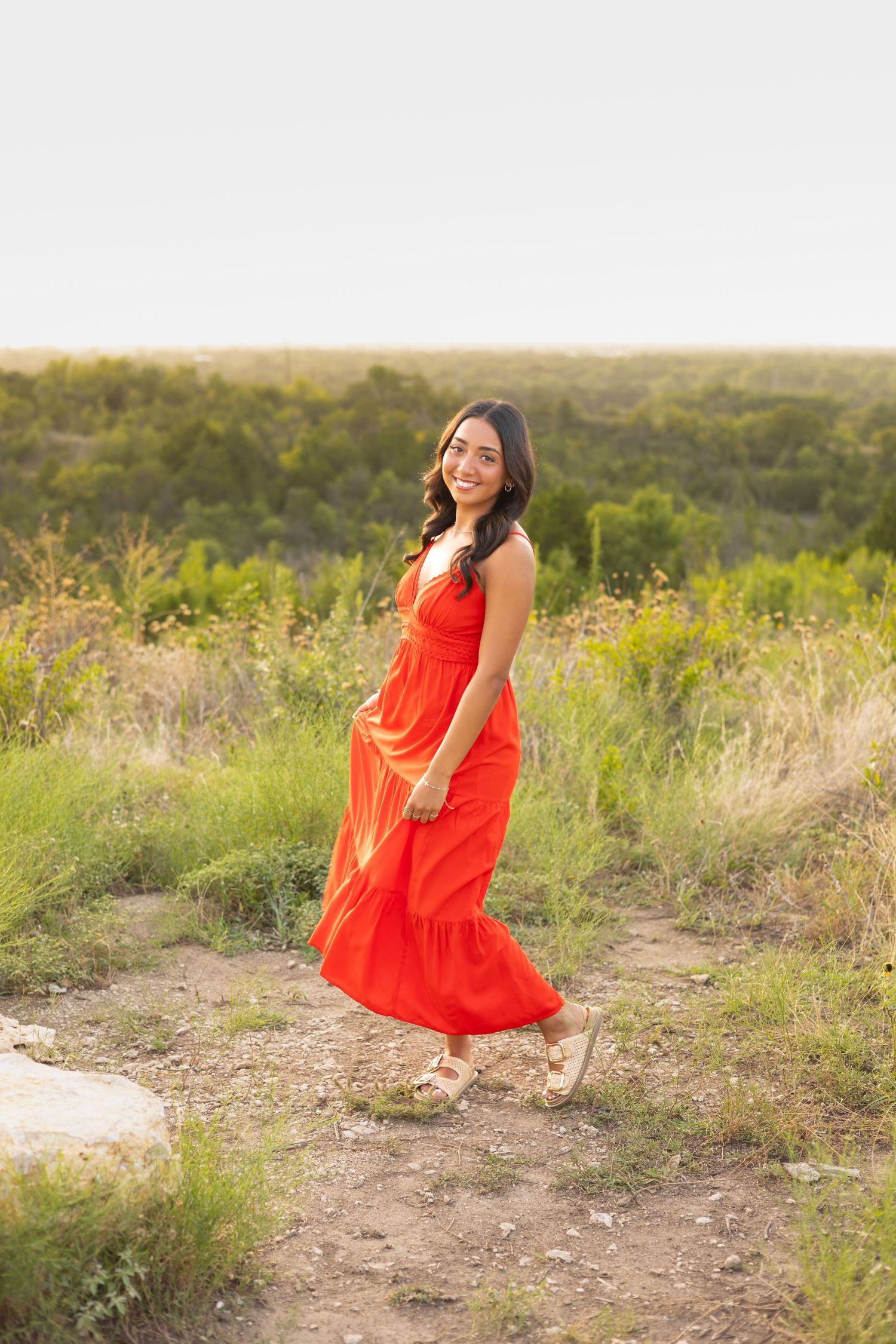 Happy woman in red maxi dress walking on a rocky trail with lush green fields behind her.