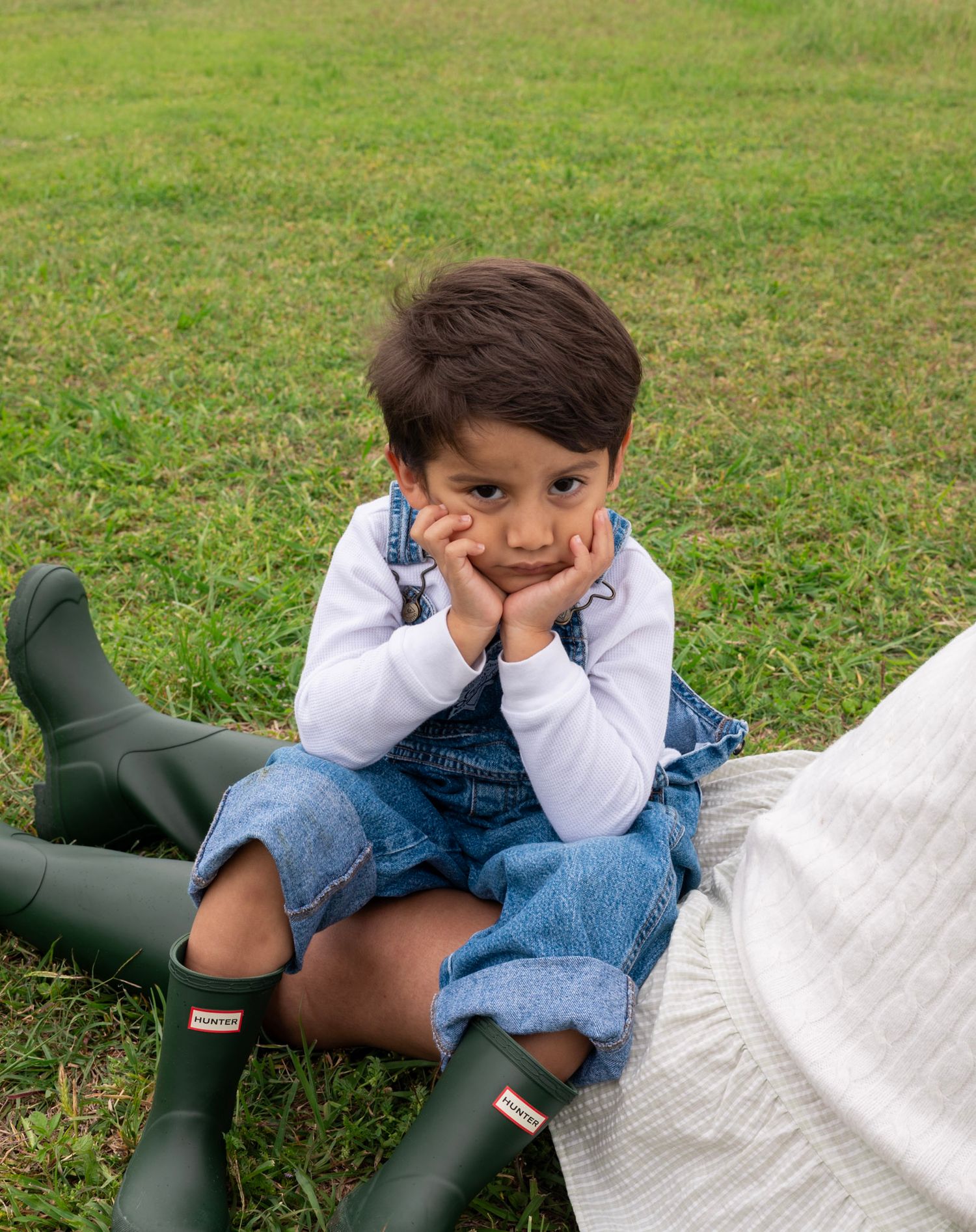 Young child in denim overalls and green Hunter boots sitting on grass with chin resting on hands.