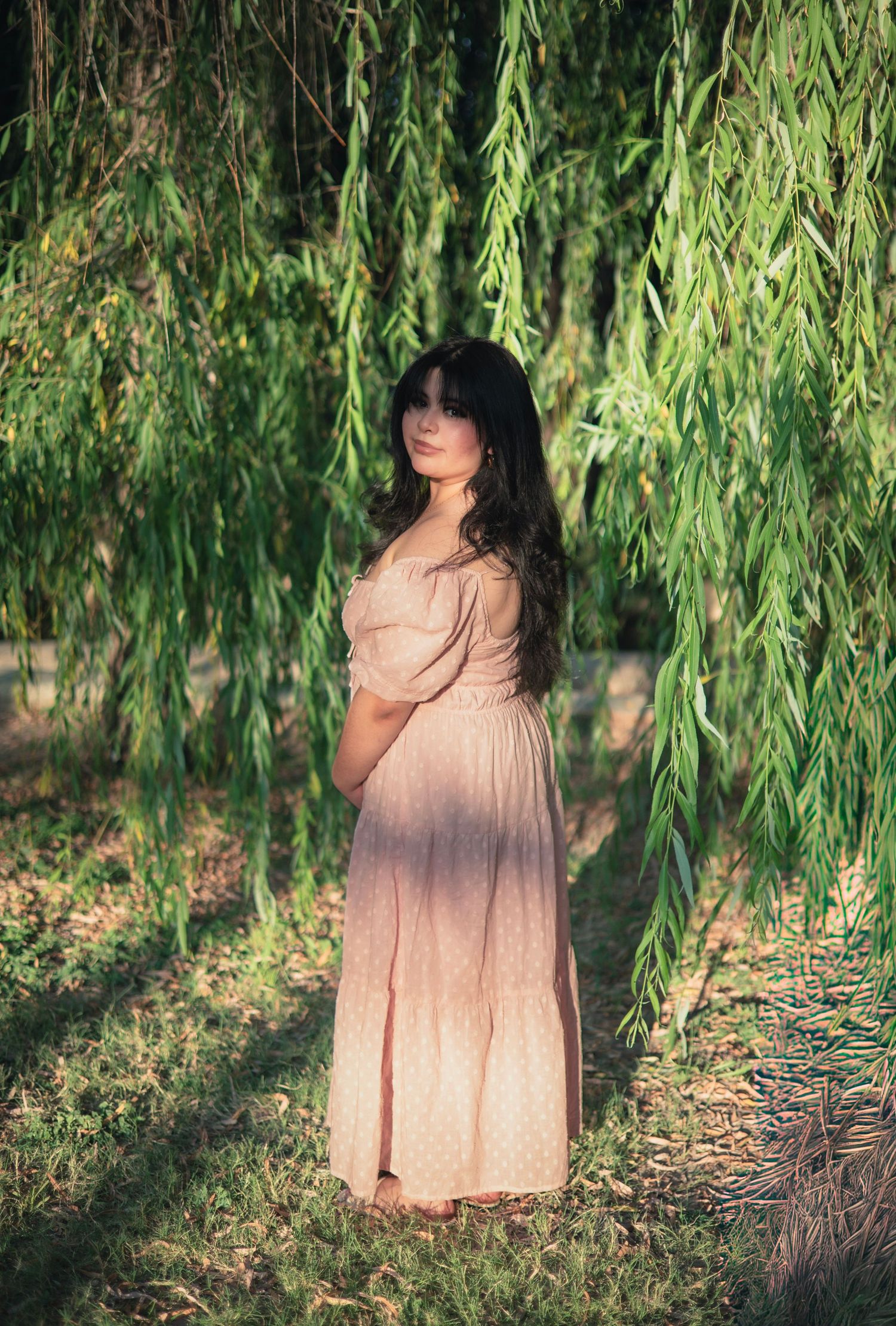 Pregnant woman in pink maxi dress standing under weeping willow tree branches in a serene outdoor setting.
