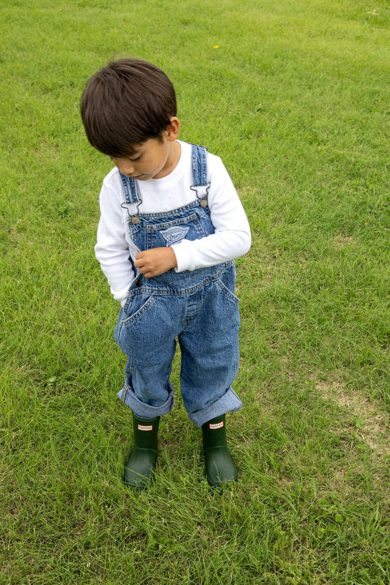 Young boy in denim overalls and green rain boots looking down on green grass.