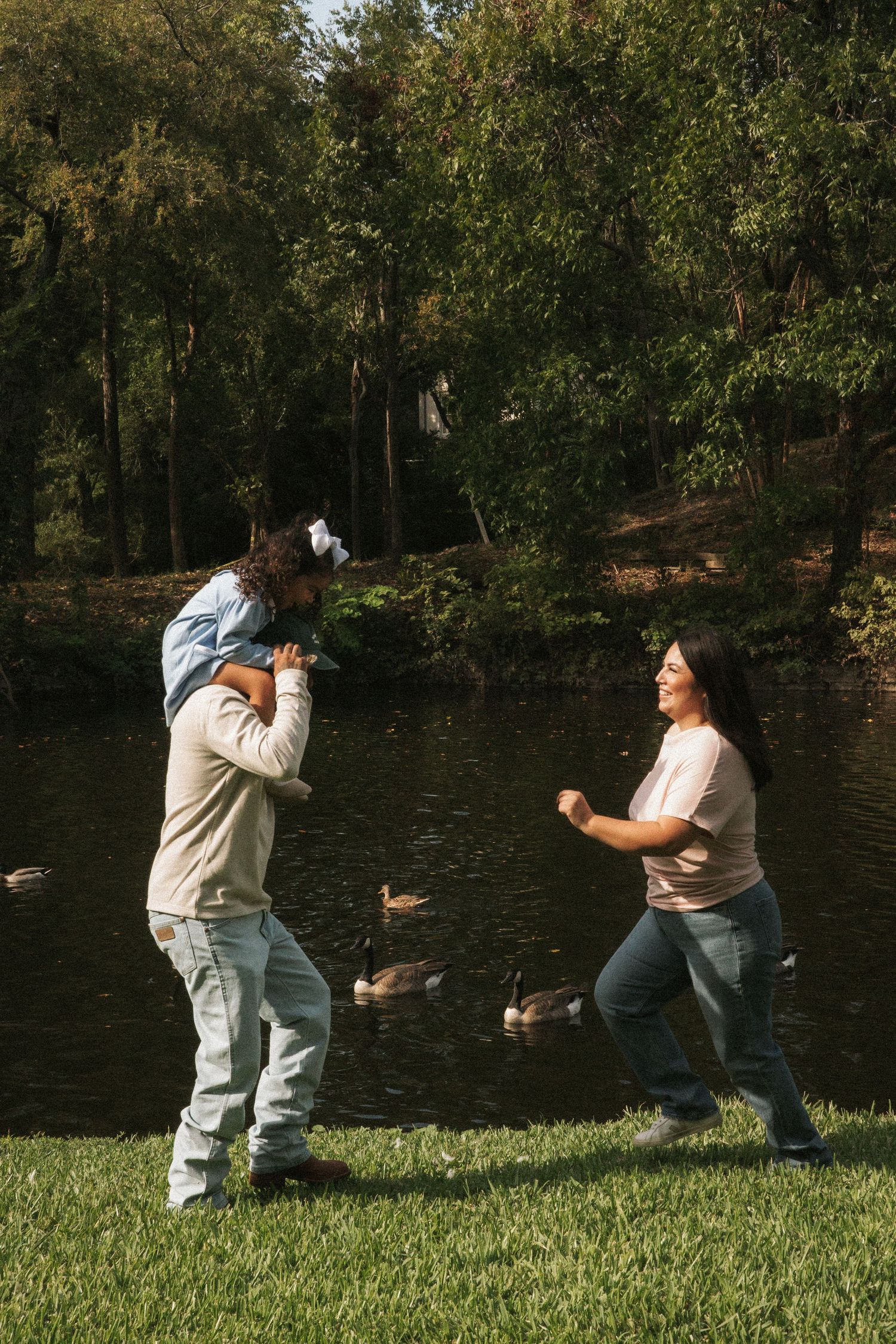 Family playing by a pond in a park, father carrying child on shoulders while mother dances nearby.