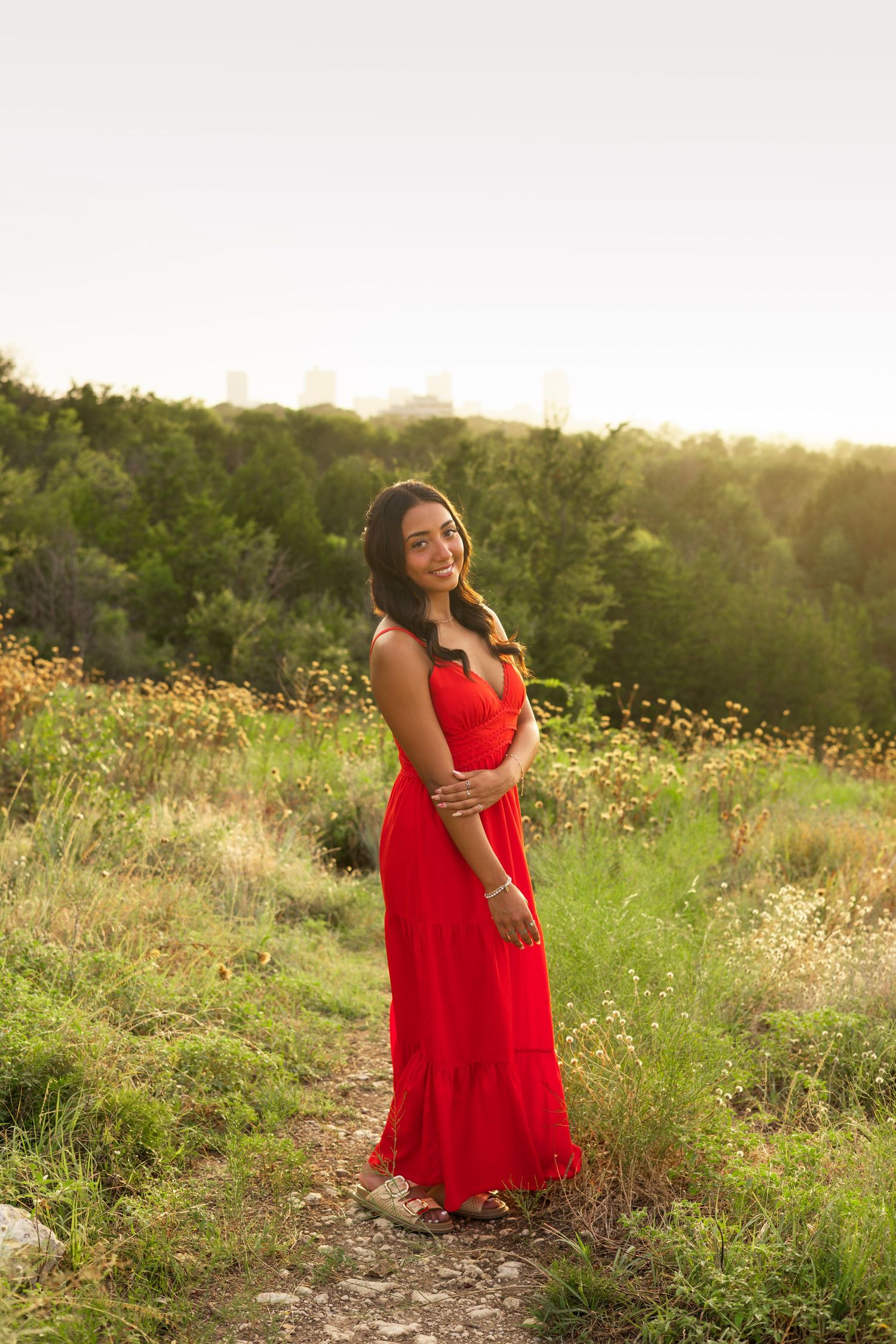 Young woman in red maxi dress smiling softly in a green meadow with city skyline behind.