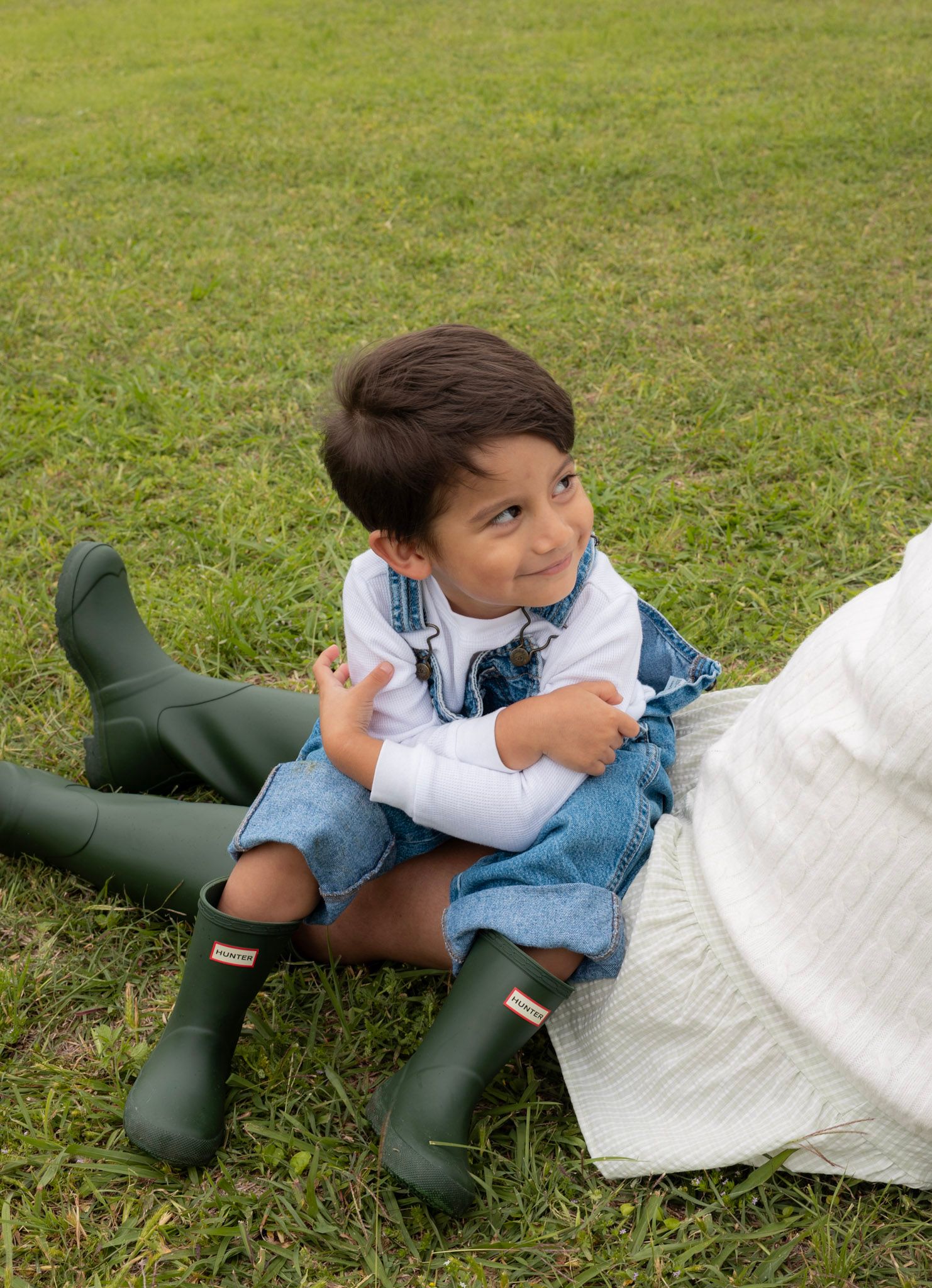 Happy toddler in denim overalls and green Hunter boots playing on grass outdoors.