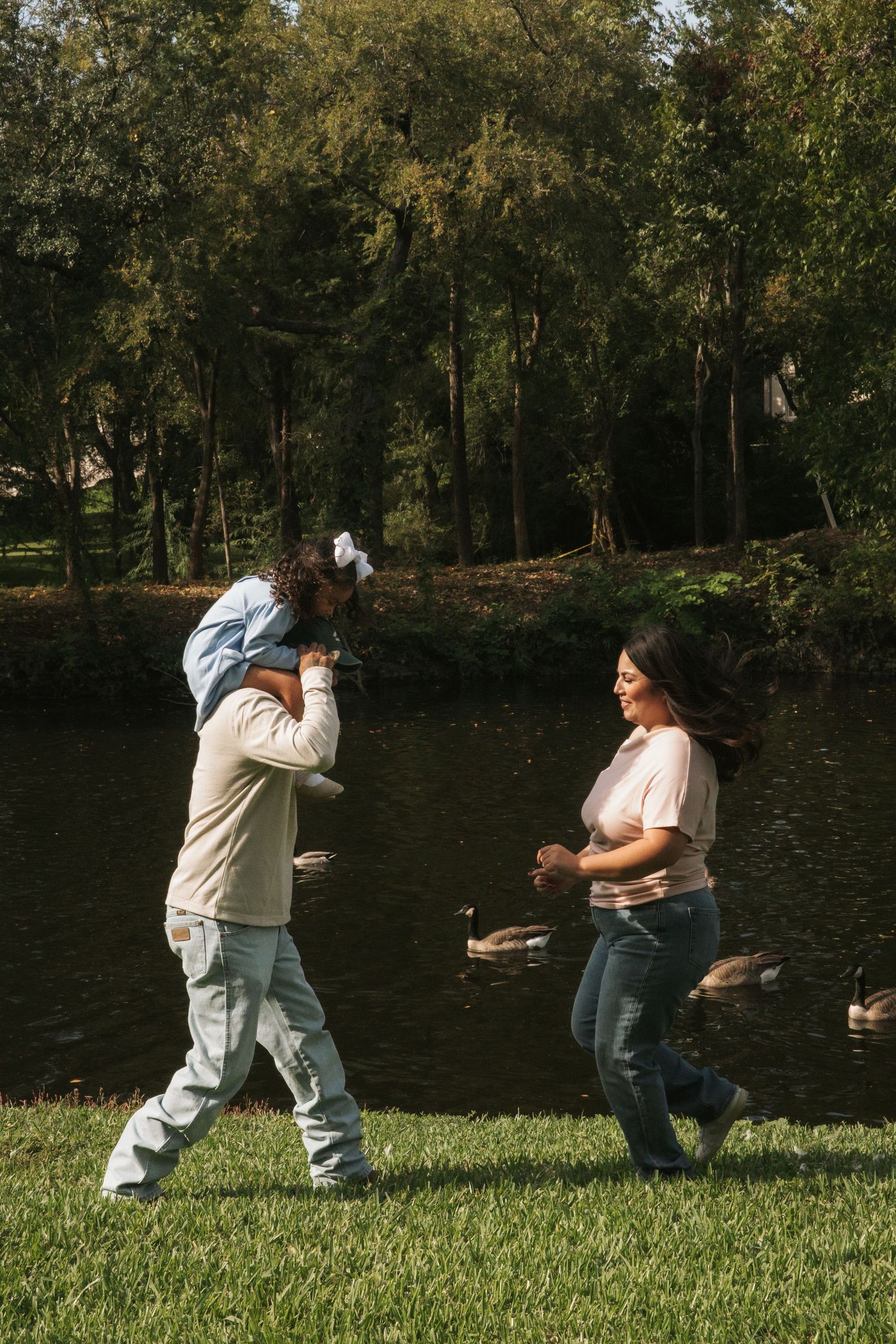 A man carries a child on his shoulders while walking with a woman by a pond in a lush green park.
