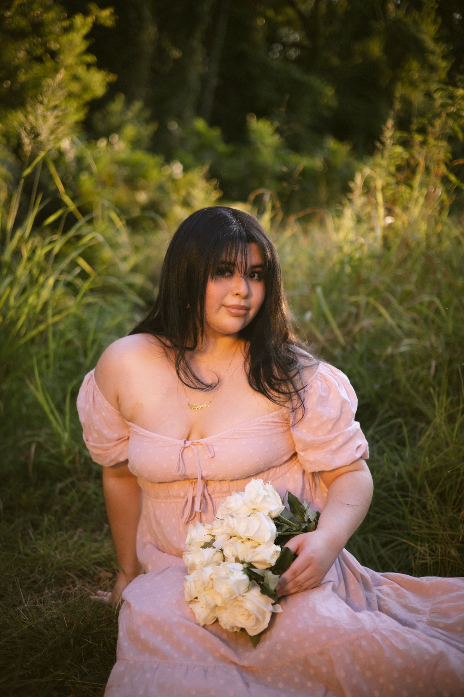 Young woman in pink off-shoulder dress sitting in grass holding white roses bouquet at sunset.