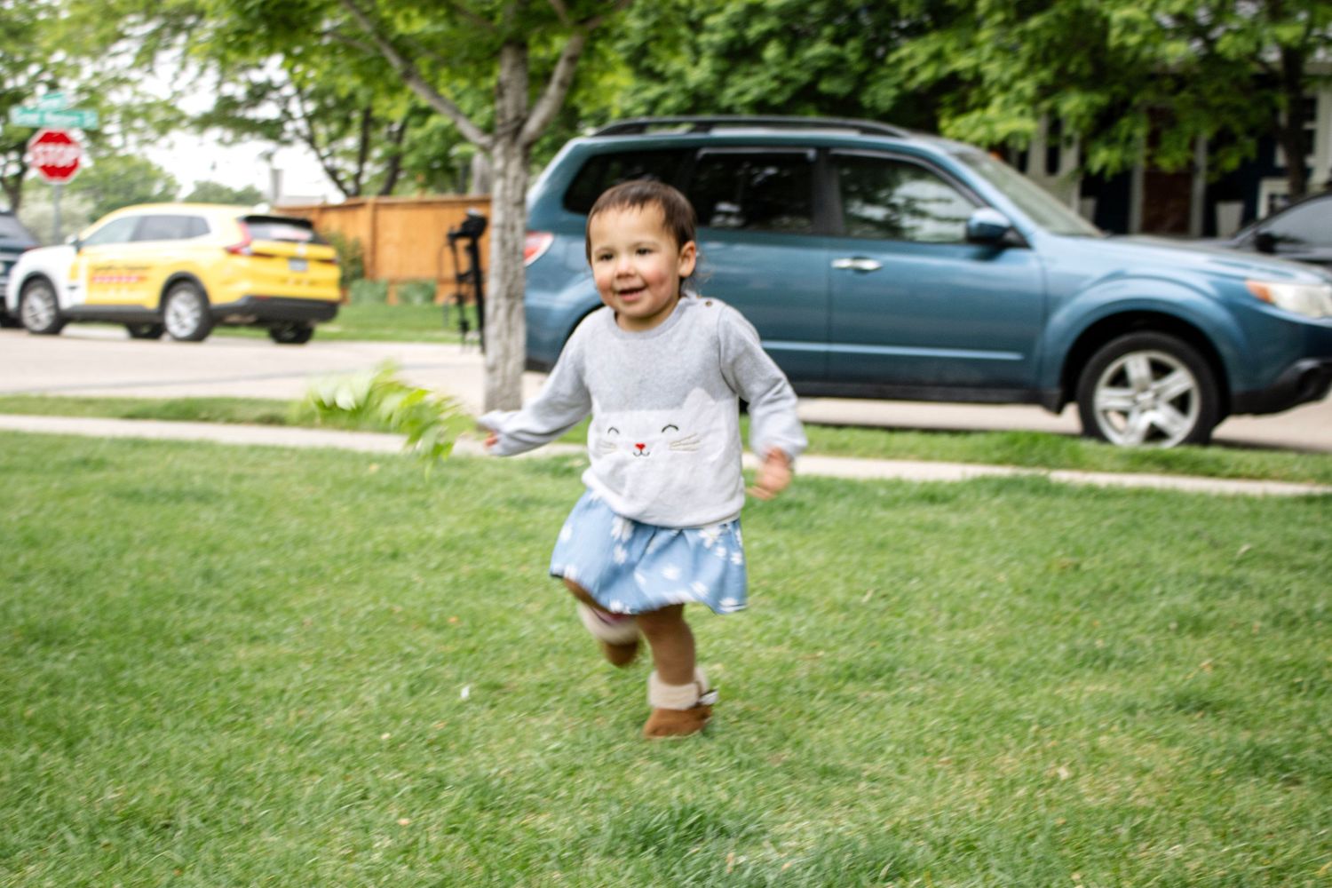 Happy toddler girl in blue skirt running on green grass in residential neighborhood.