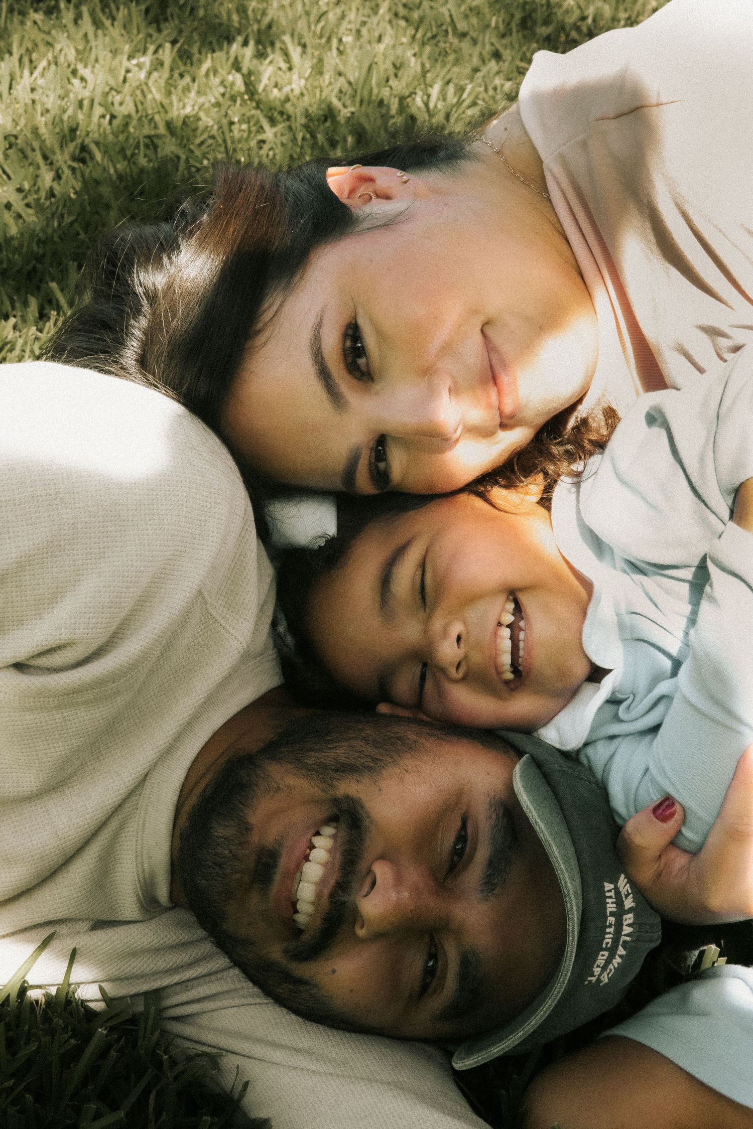Happy family of three lying on grass, smiling together in a warm outdoor portrait.