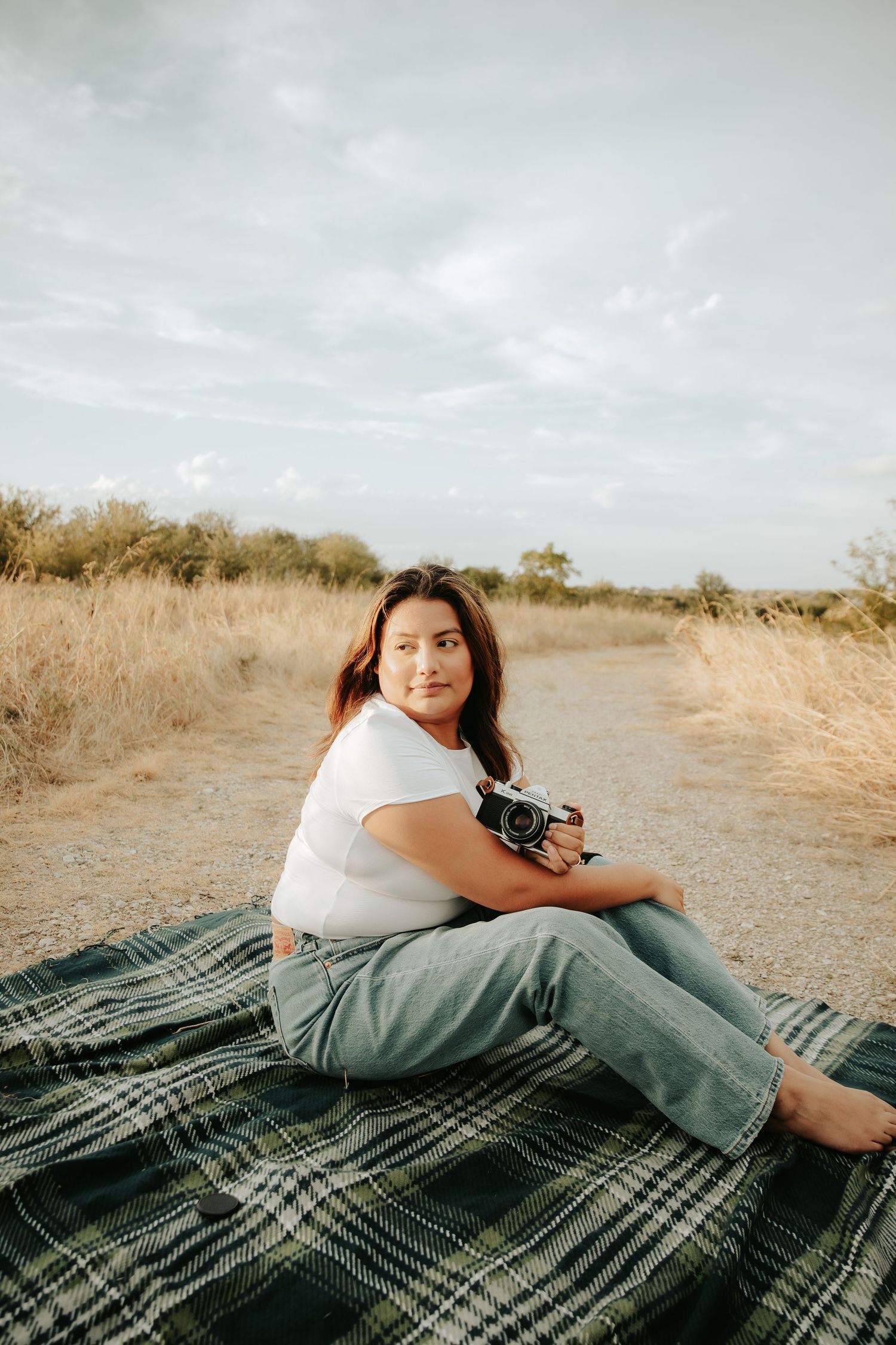 Woman sitting on plaid blanket outdoors holding vintage camera, wearing white tee and jeans, with dry grassland background.