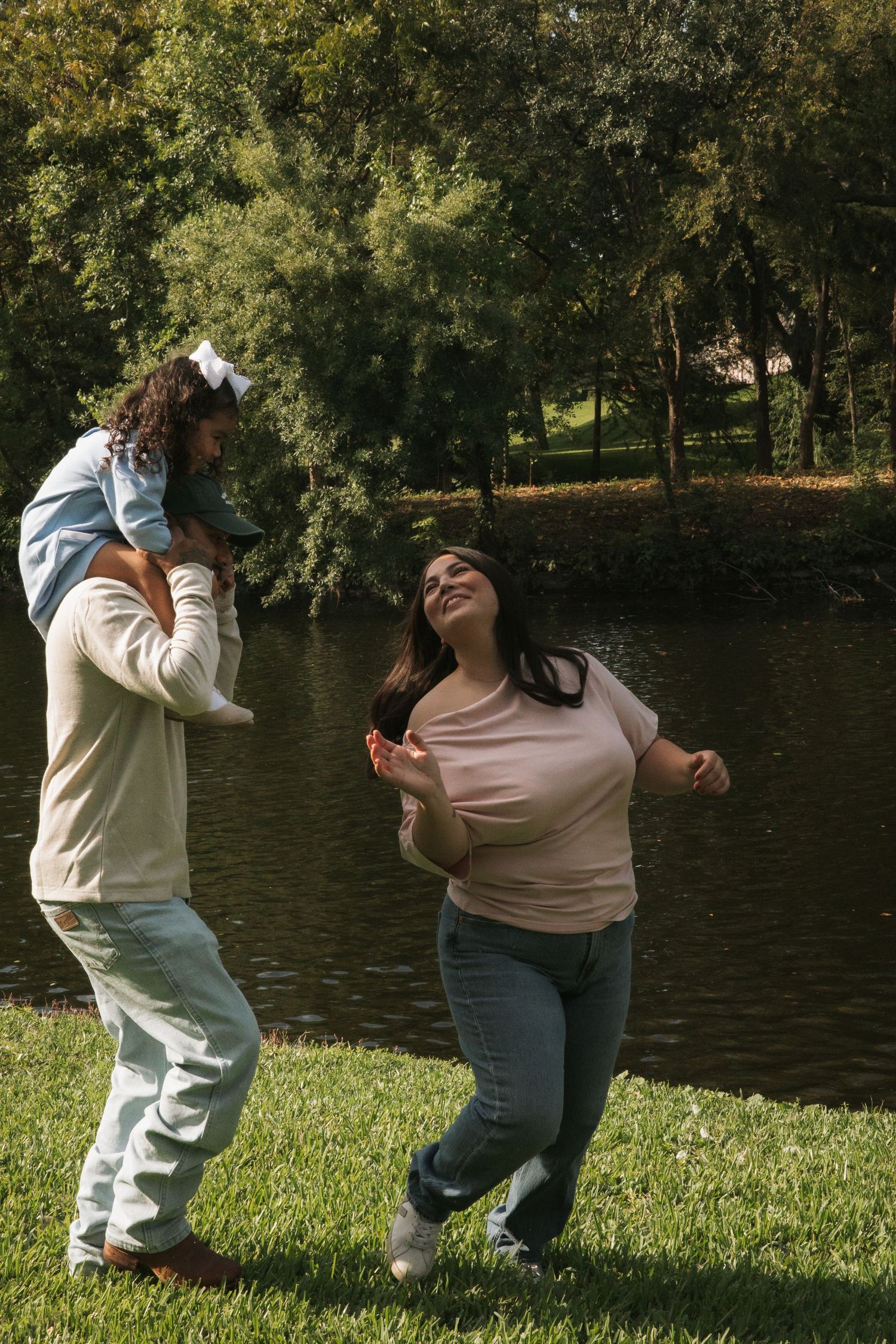 Joyful family moment by a pond, father carrying toddler on shoulders while mother dances.