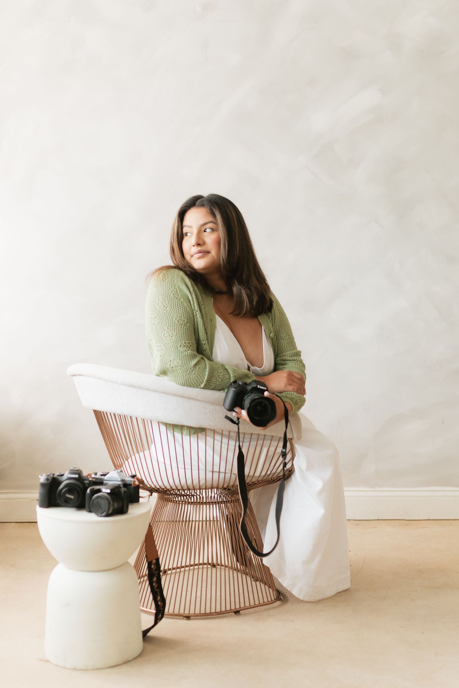 Female photographer in green cardigan sitting in chair gazing away holding camera indoors.