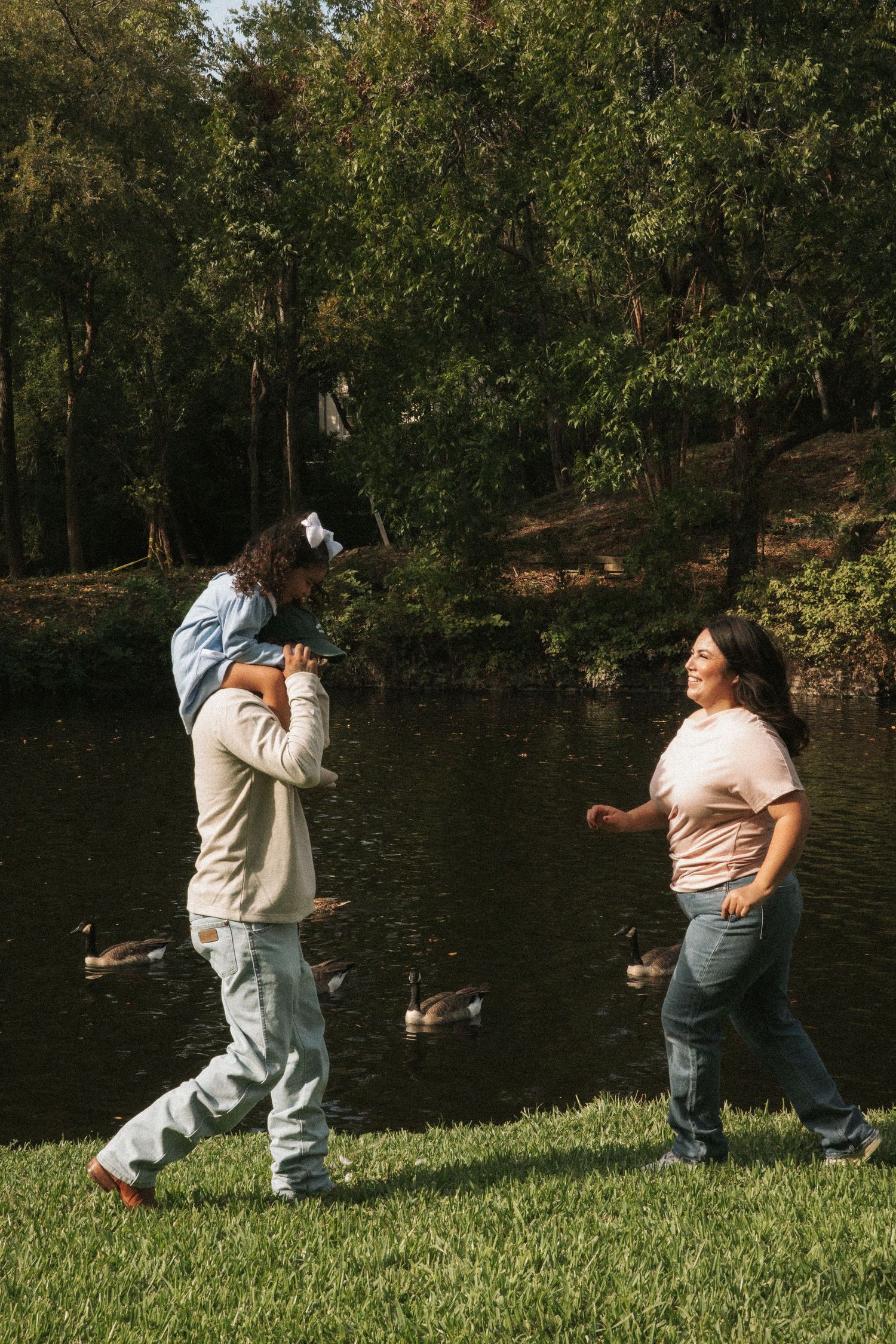 Family enjoying a sunny day by a pond, with a child on dad's shoulders and mom walking alongside ducks.