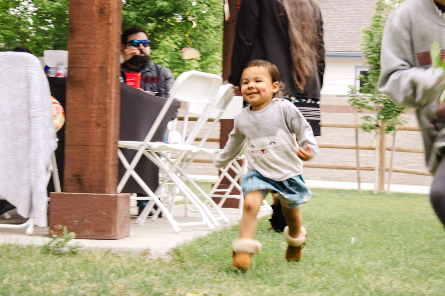 A joyful toddler runs across a green lawn at an outdoor gathering, wearing a gray shirt and blue skirt.