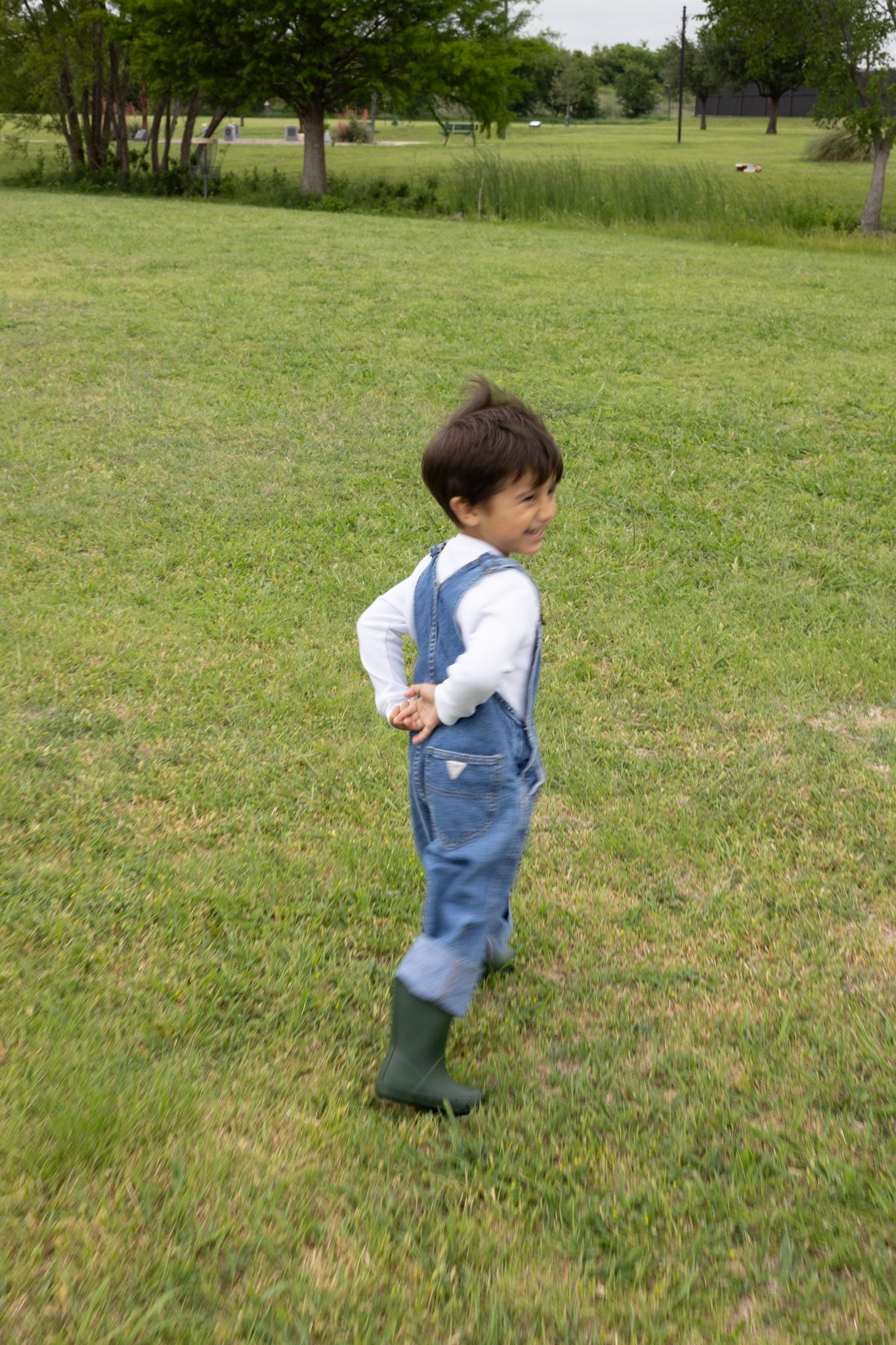 Happy young toddler in denim overalls and green rain boots running and smiling in a park.