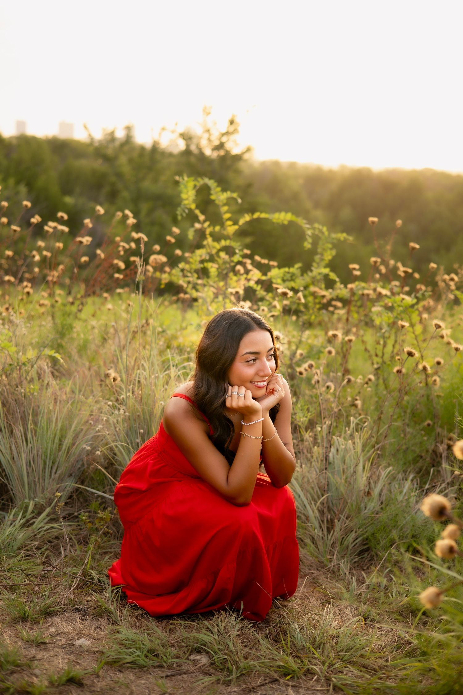 Smiling young woman in red dress sitting among wildflowers in a sun-drenched natural meadow.