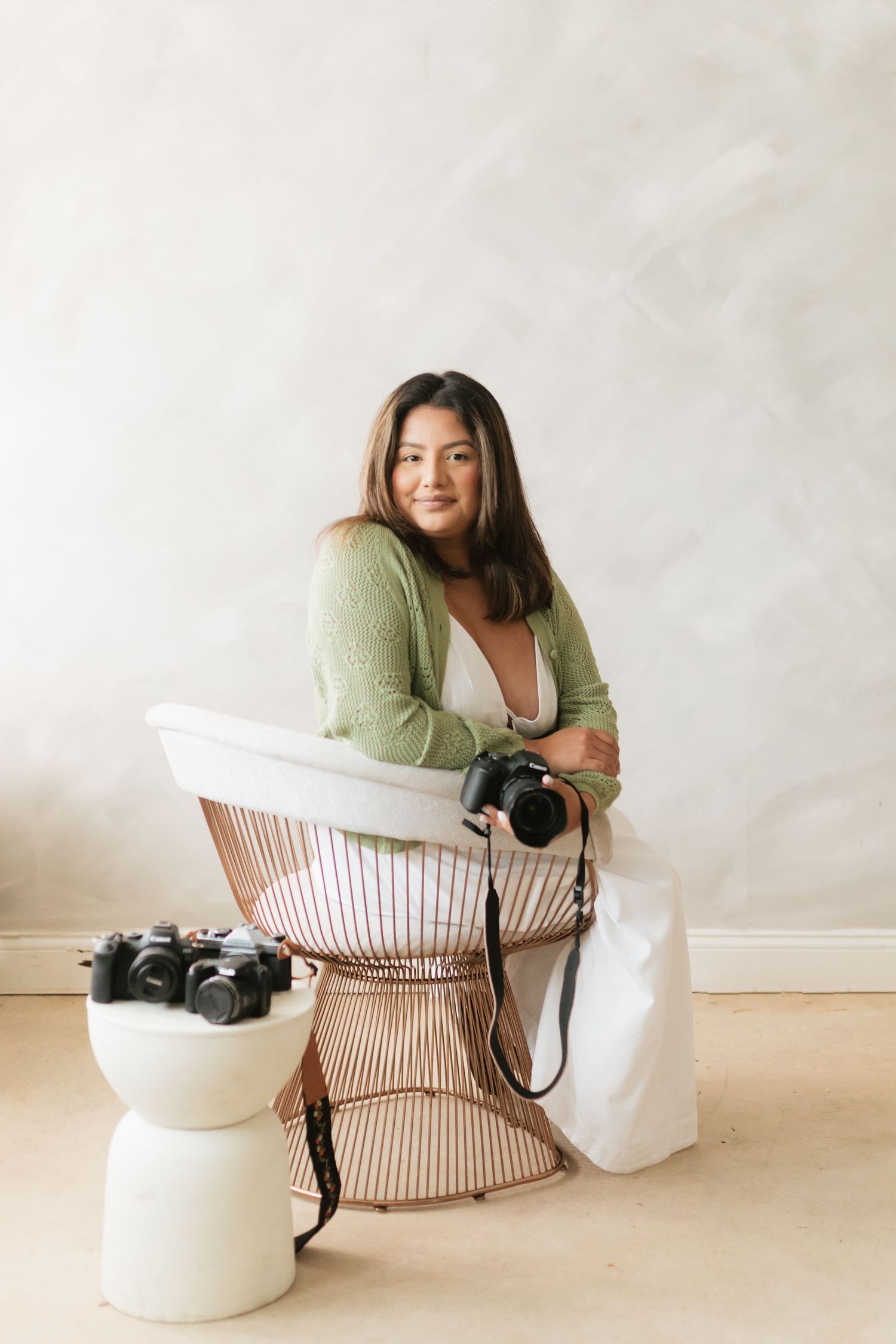 Female photographer in green cardigan sitting in chair holding camera with cameras nearby.