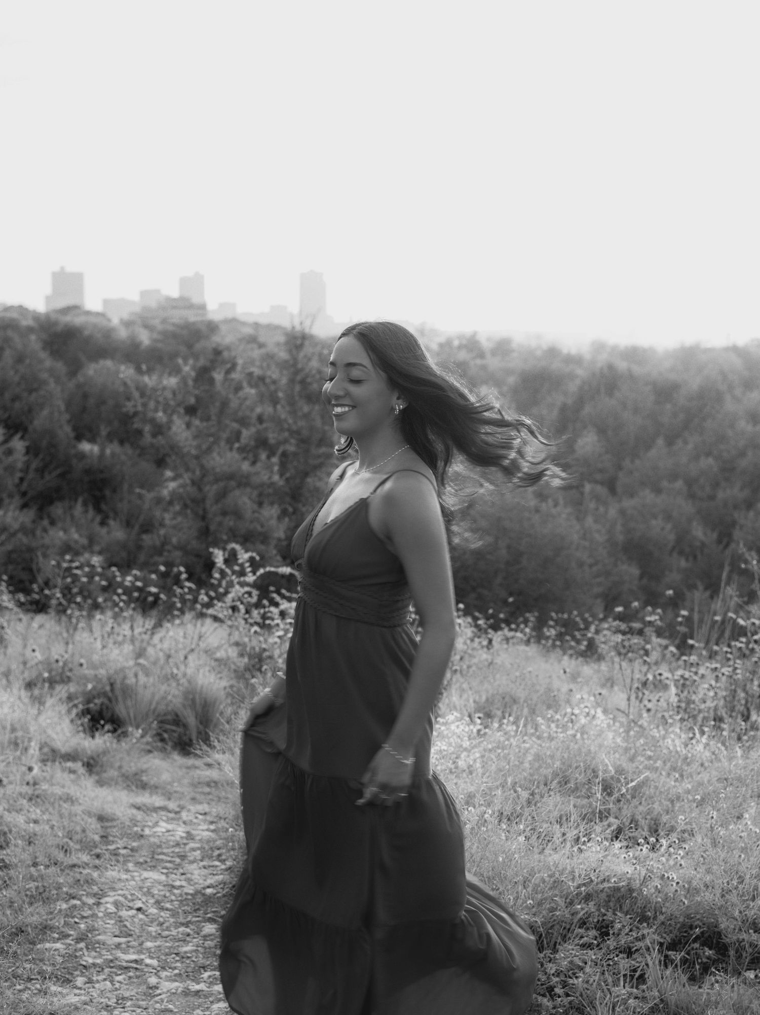 Young woman in flowing dress twirling on a hilltop trail with city skyline in black and white.