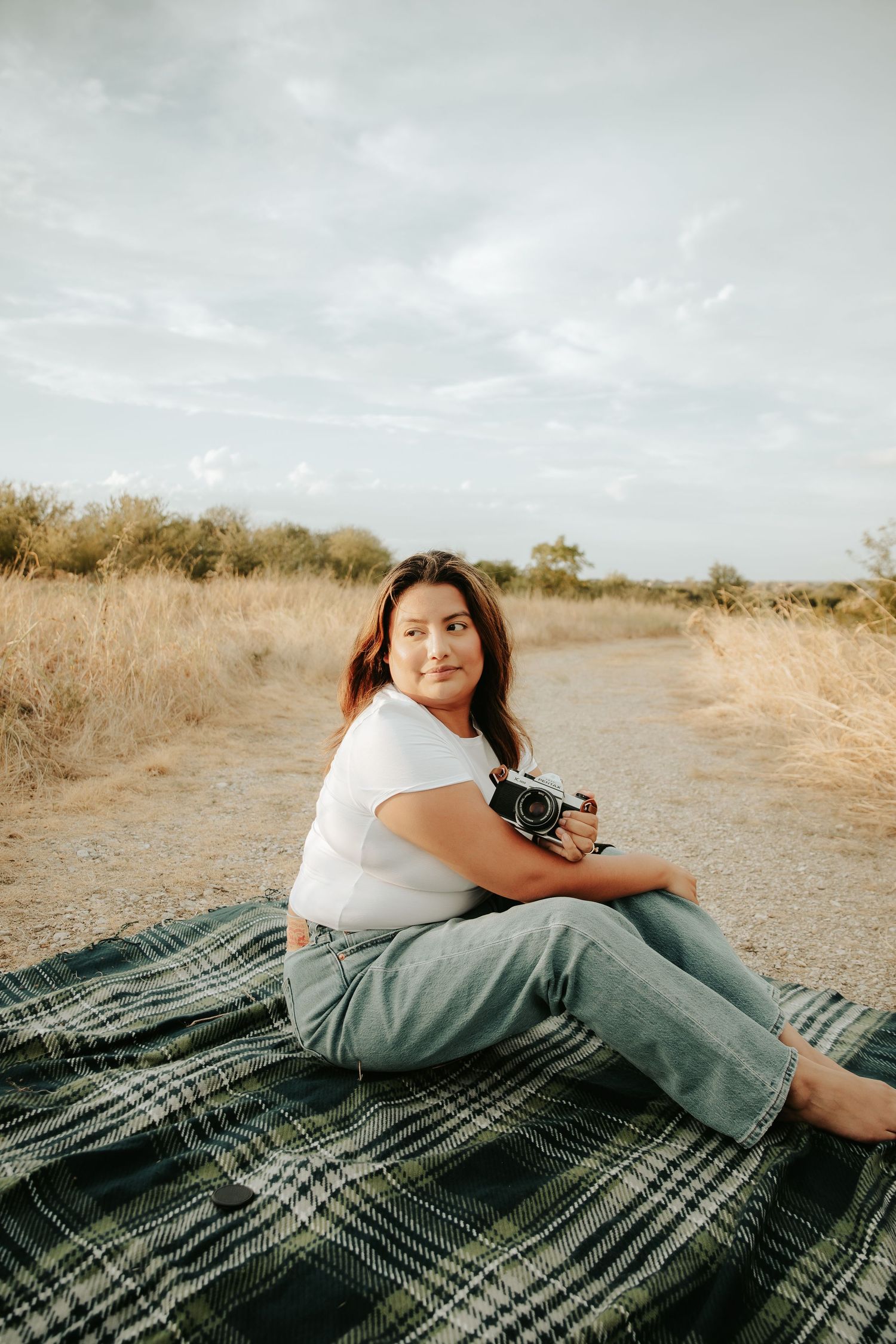 Woman sitting on plaid blanket outdoors holding vintage camera, smiling in natural field setting.