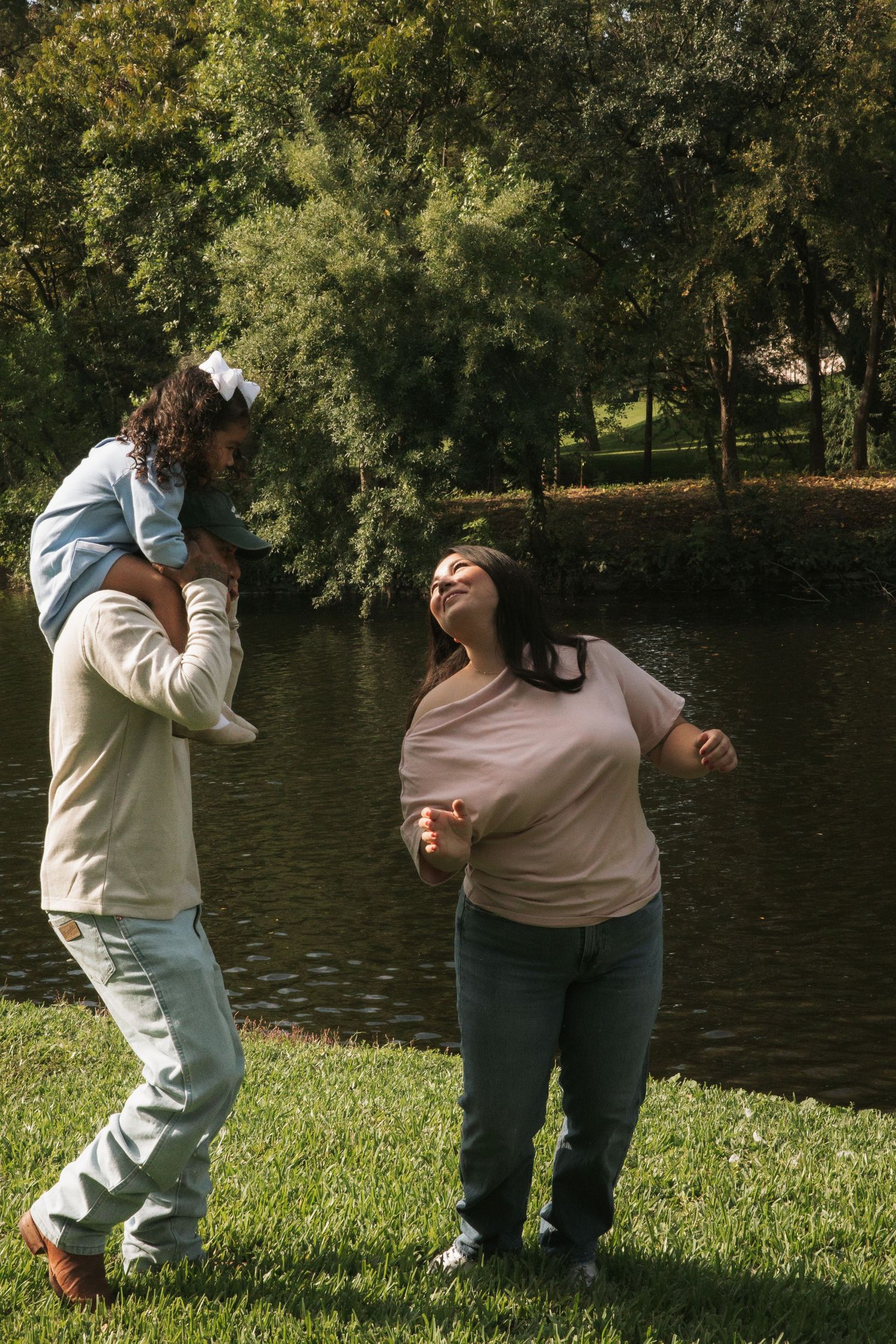Joyful family moment as father lifts young daughter on shoulders while mother laughs outdoors by pond.